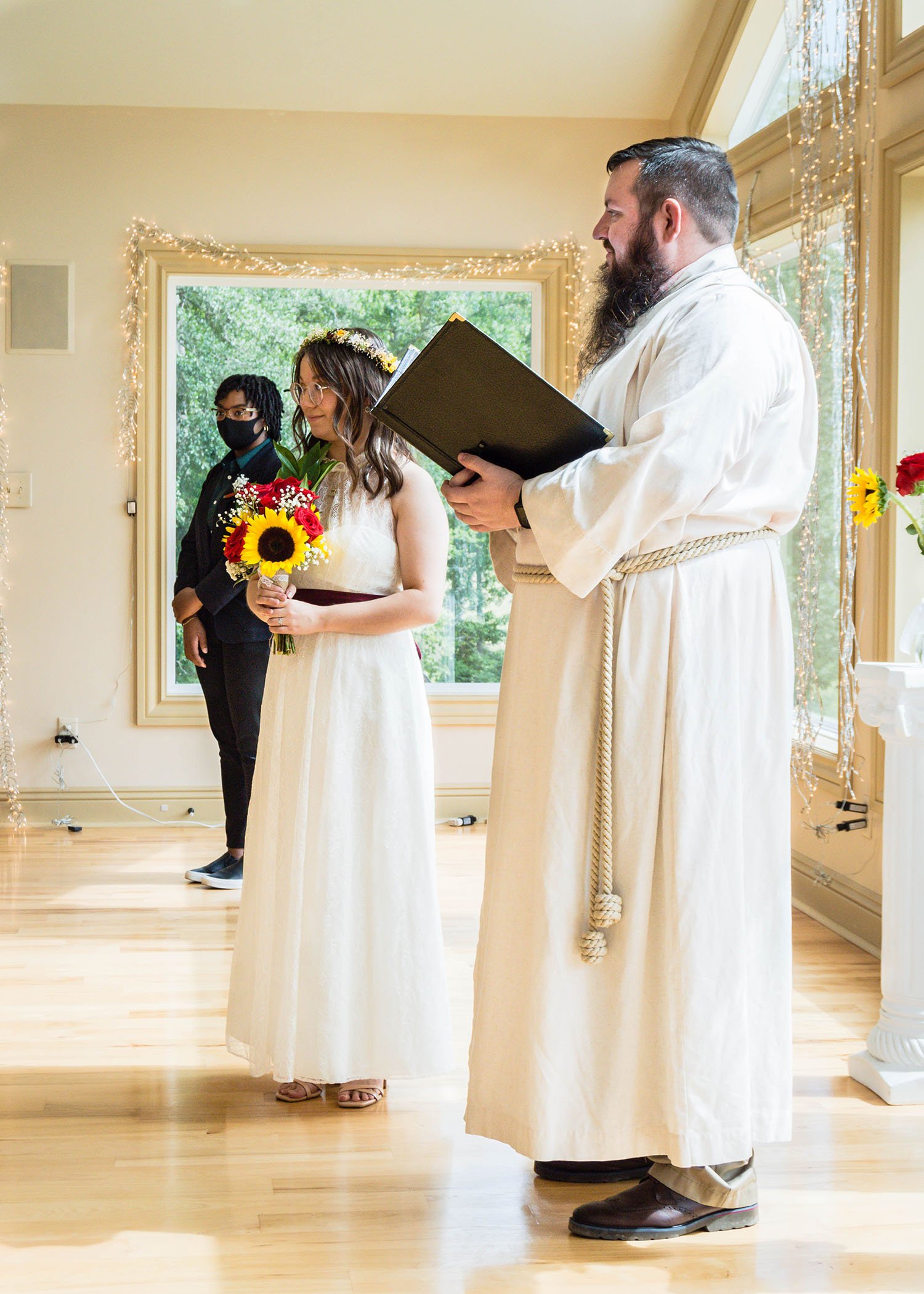 An LGBTQ+ marrier stands between their officiant and wedding party as they wait patiently for their partner to walk down the aisle.