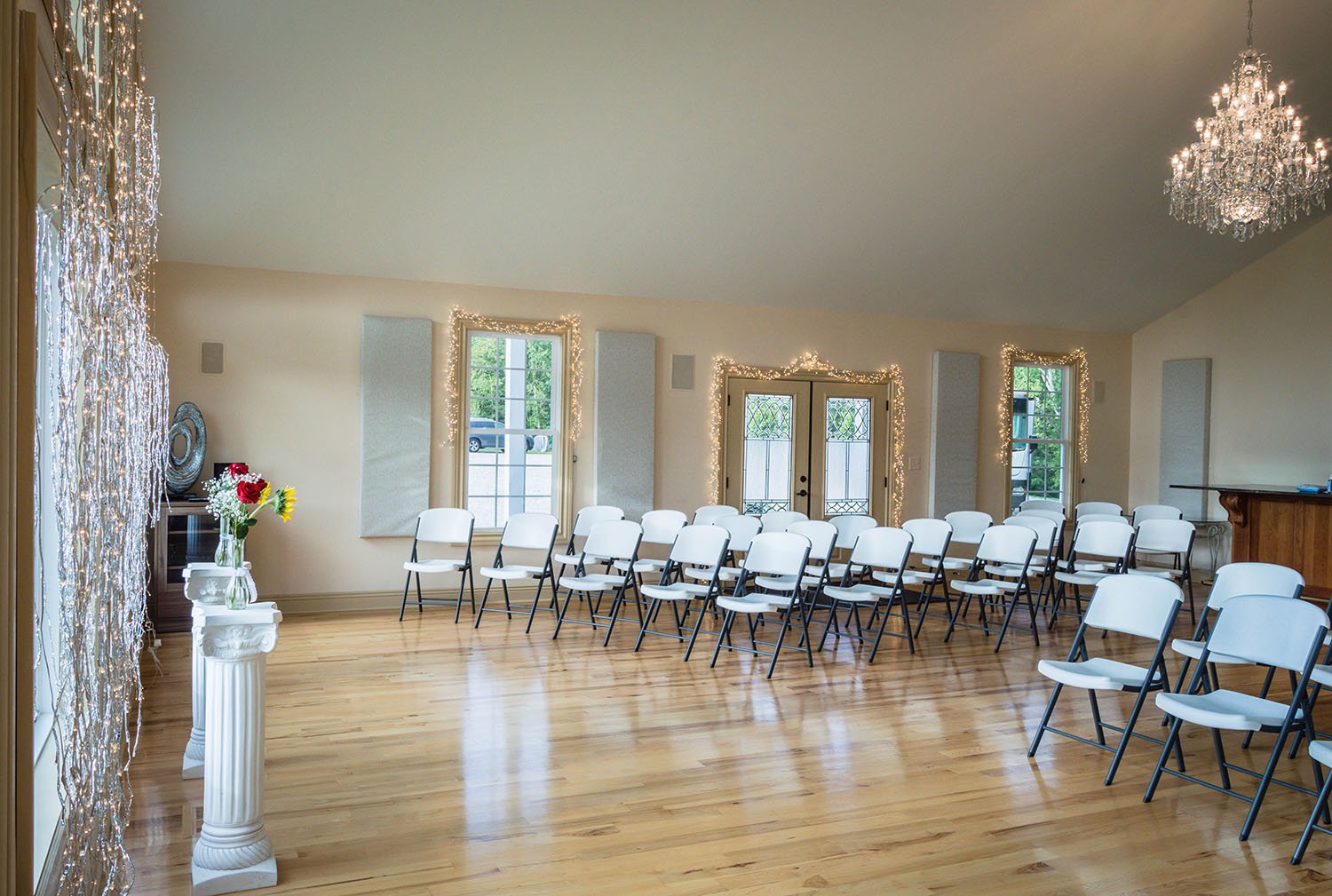 Empty white chairs are set up in the wedding venue and, at the altar, there are two pillars with vases with sunflowers and roses.