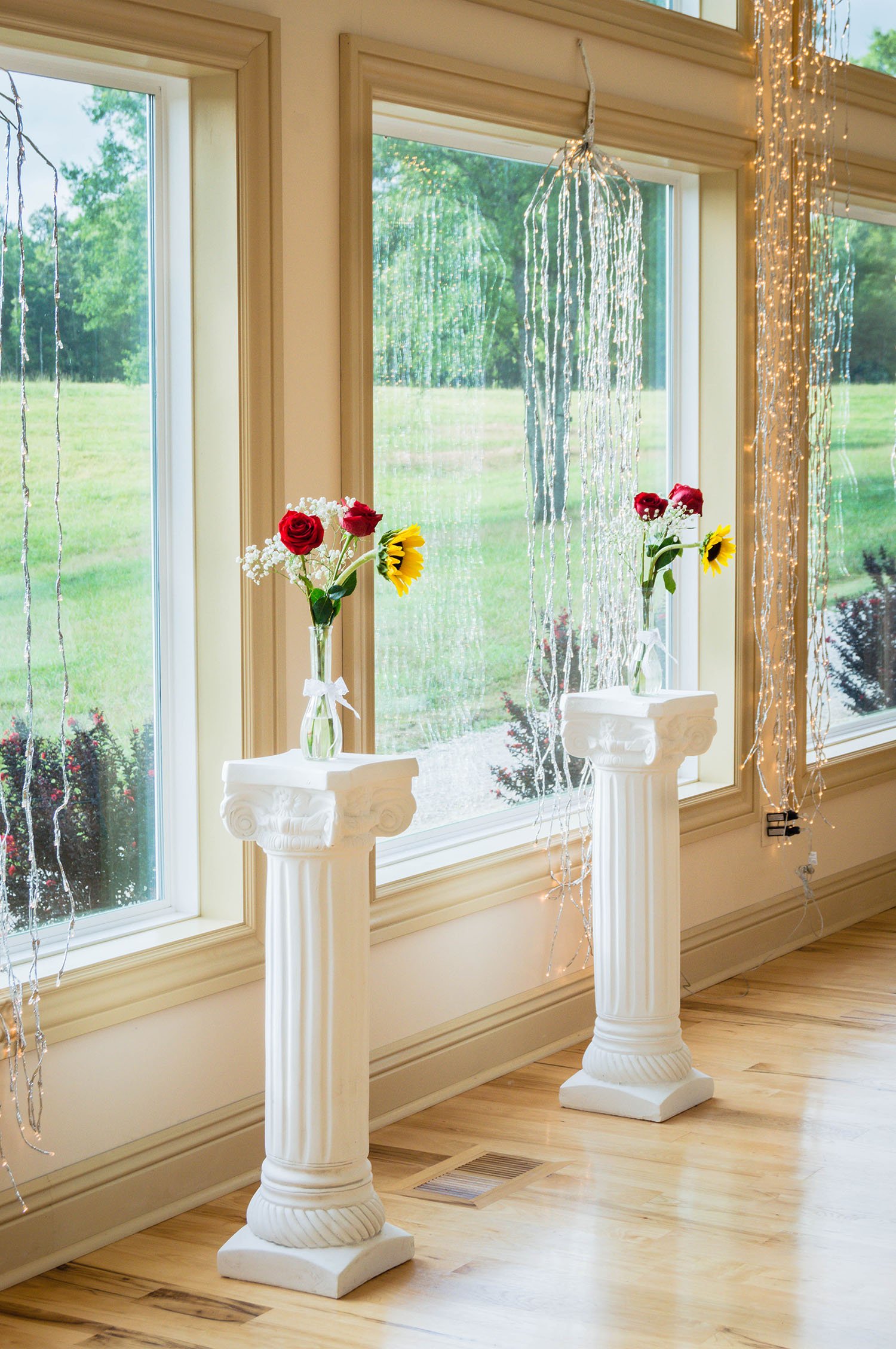 At the alter are two white pillars with small vases filled with water, roses, sunflowers, and baby's breath flower arrangements with the string lights draping over the large windows behind them.