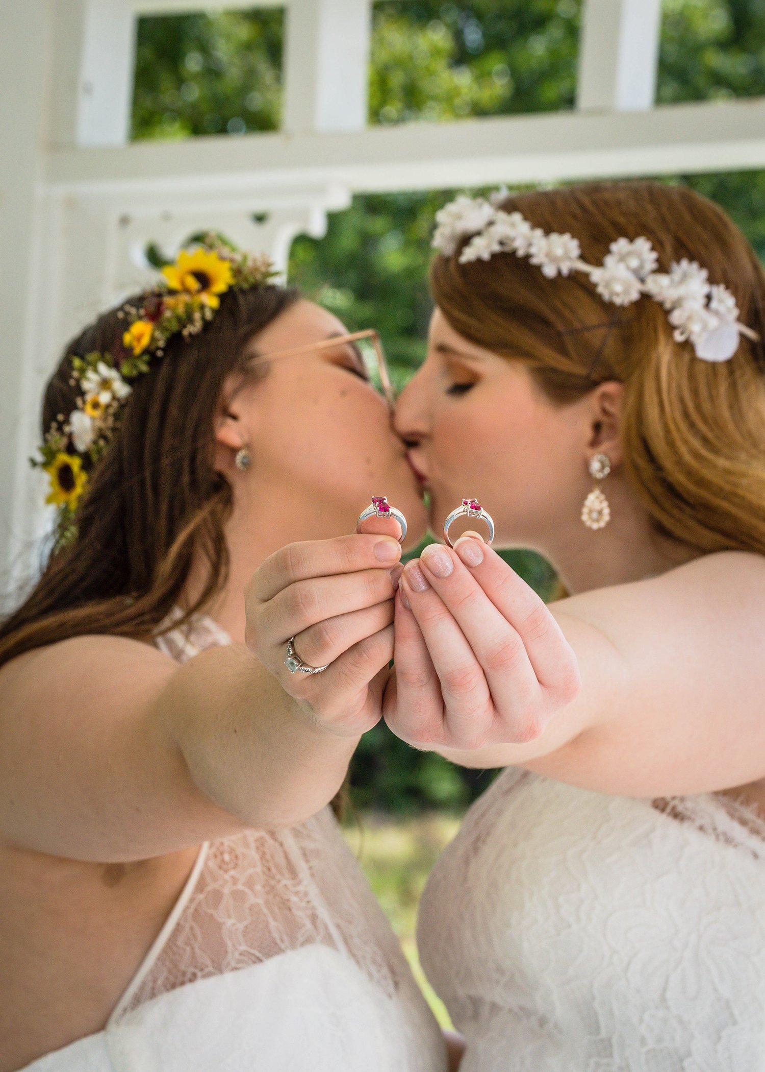 Two LGBTQ+ brides kiss one another as they hold out their wedding rings.