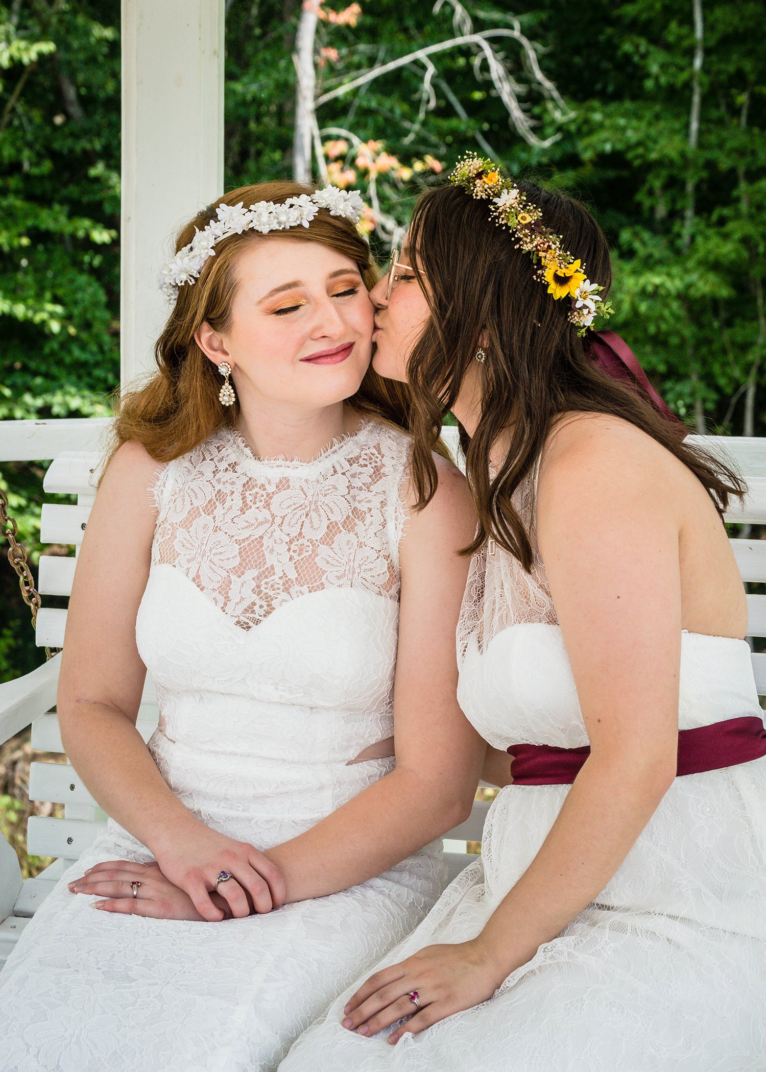 Two LGBTQ+ marriers sit on a bench in a gazebo on the wedding venue. One marrier kisses the cheek of their partner while the partner smiles with eyes closed.