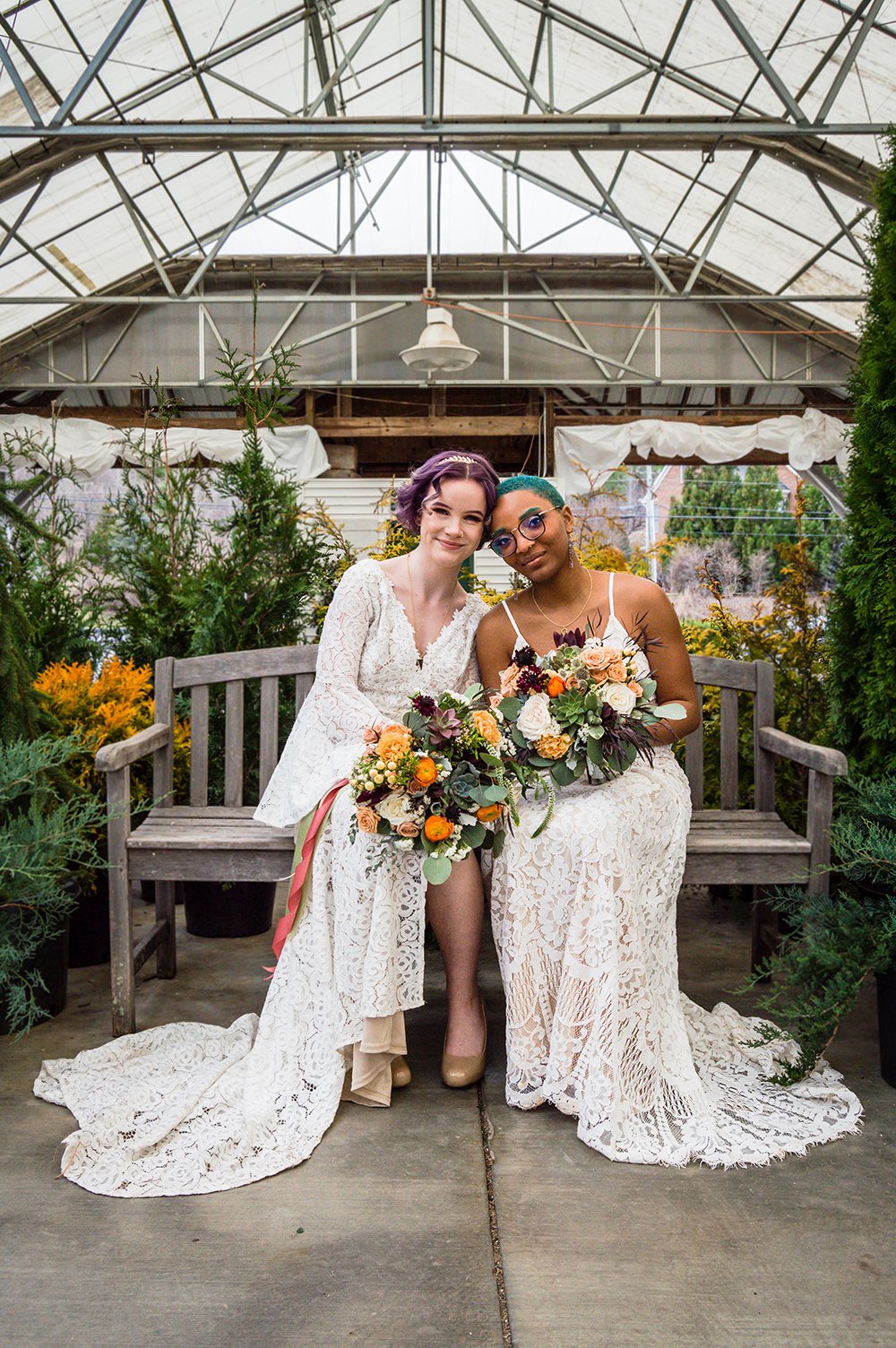 An LGBTQ+ wedding couple sit on a bench, hold their bouquets, and smile for a photo during their elopement.