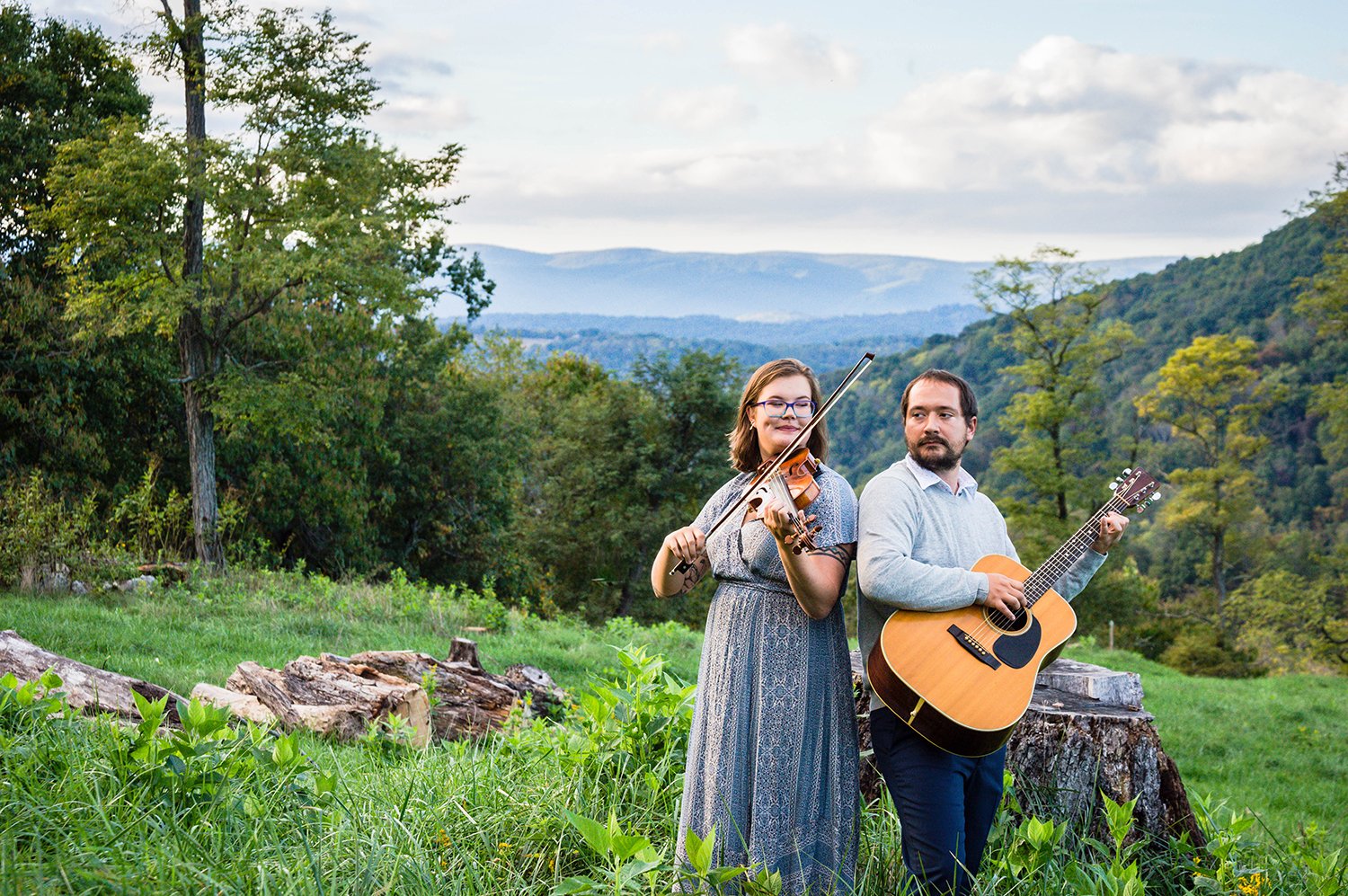 A couple plays a violin and guitar atop of a hill overlooking the Blue Ridge Mountains for a couple's adventure session in Blacksburg, Virginia.