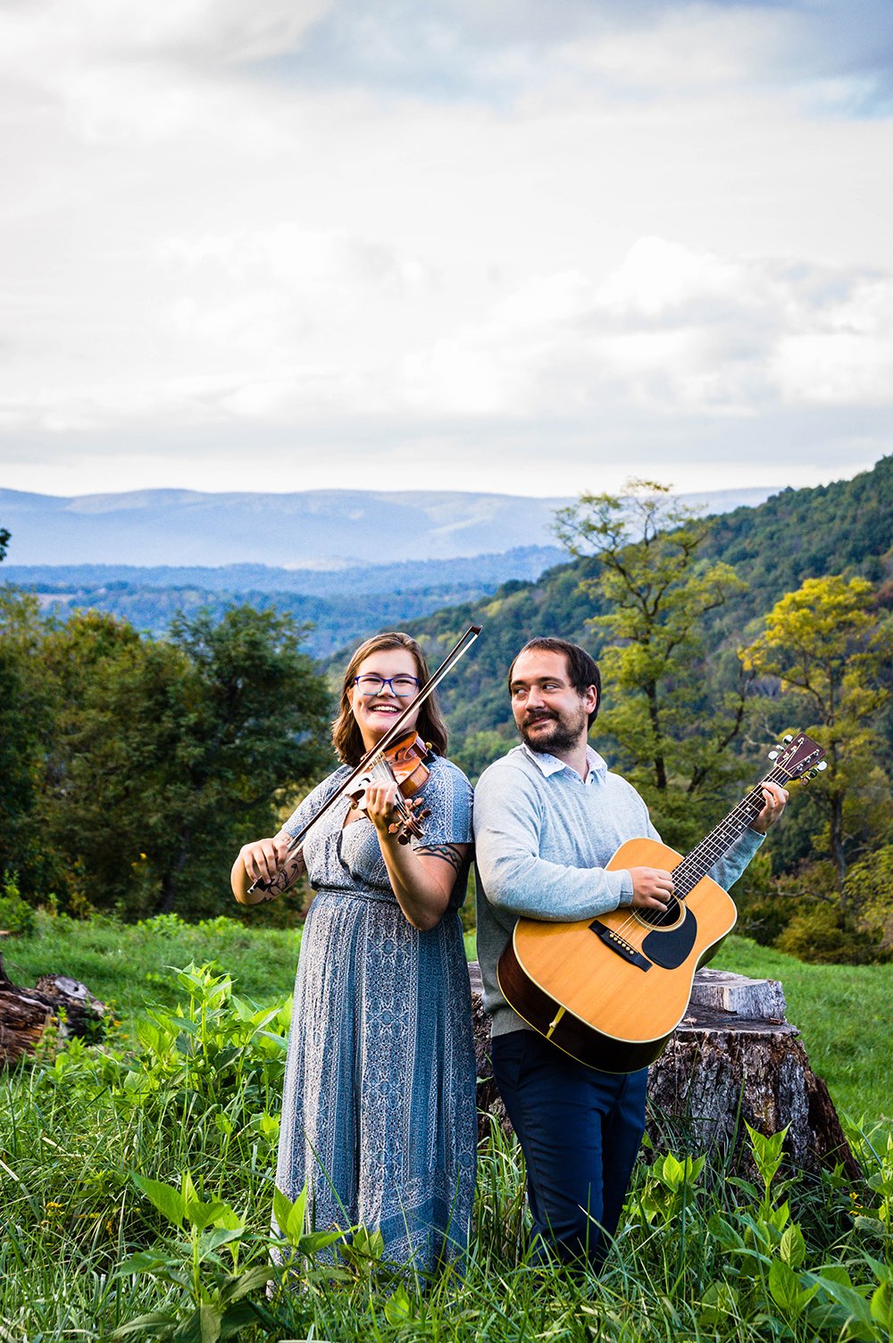 A couple plays a violin and guitar atop of a hill overlooking the Blue Ridge Mountains for a couple's adventure session in Blacksburg, Virginia.