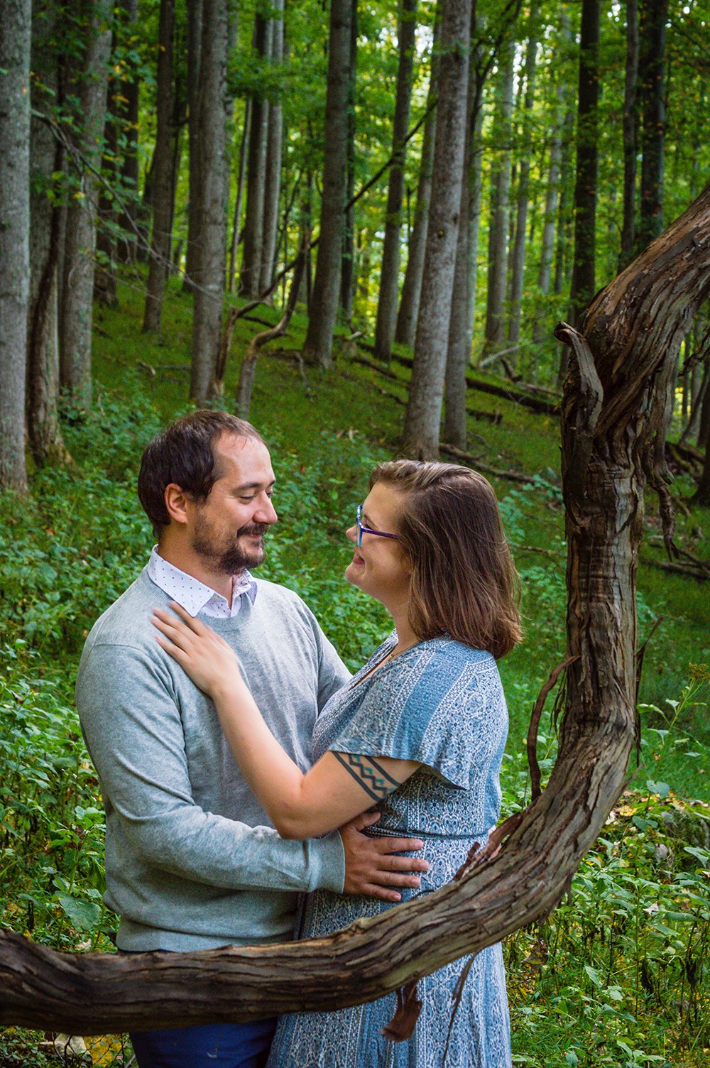 A couple embraces behind a bent branch on the bottom of a forested hill during their adventure couple’ session in Blacksburg, Virginia.