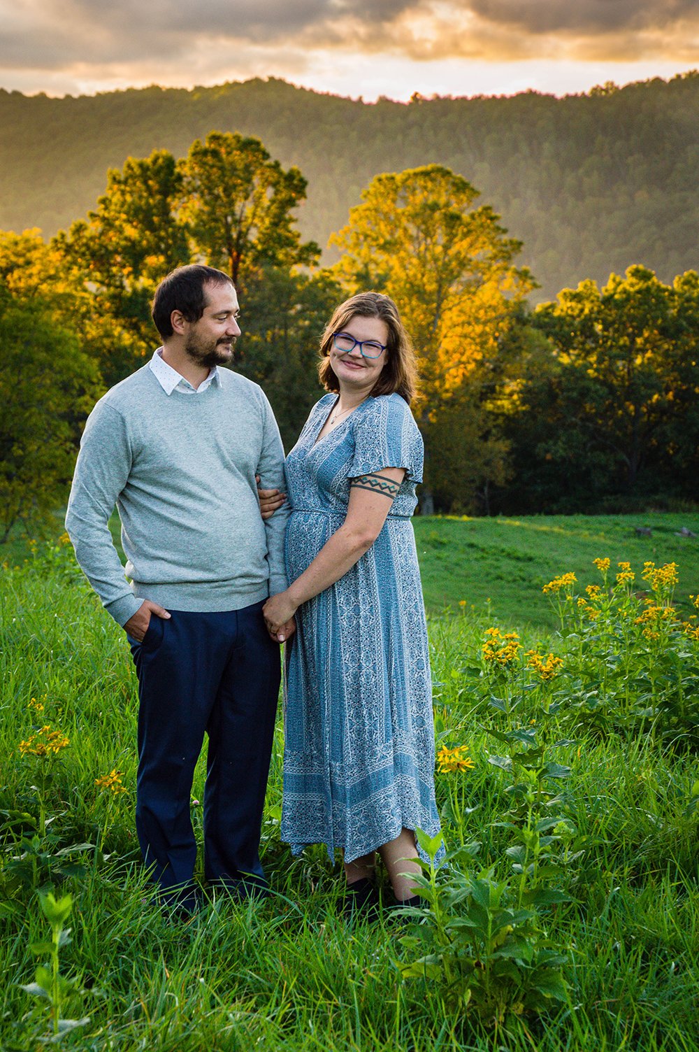 A couple stands atop of a hill overlooking the Blue Ridge Mountains with the sunset hitting against tall trees.