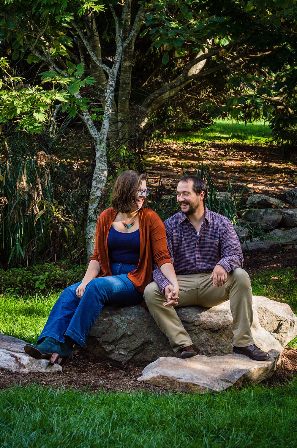 A couple sits on a large rock surrounded by various plants in the Hahn Horticulture Garden on Virginia Tech's main campus for a couple's adventure session in Blacksburg, Virginia.