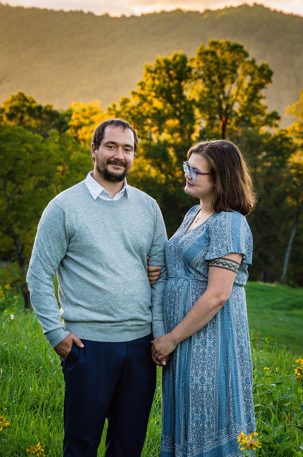 A couple stands atop of a hill overlooking the Blue Ridge Mountains with the sunset hitting against tall trees.