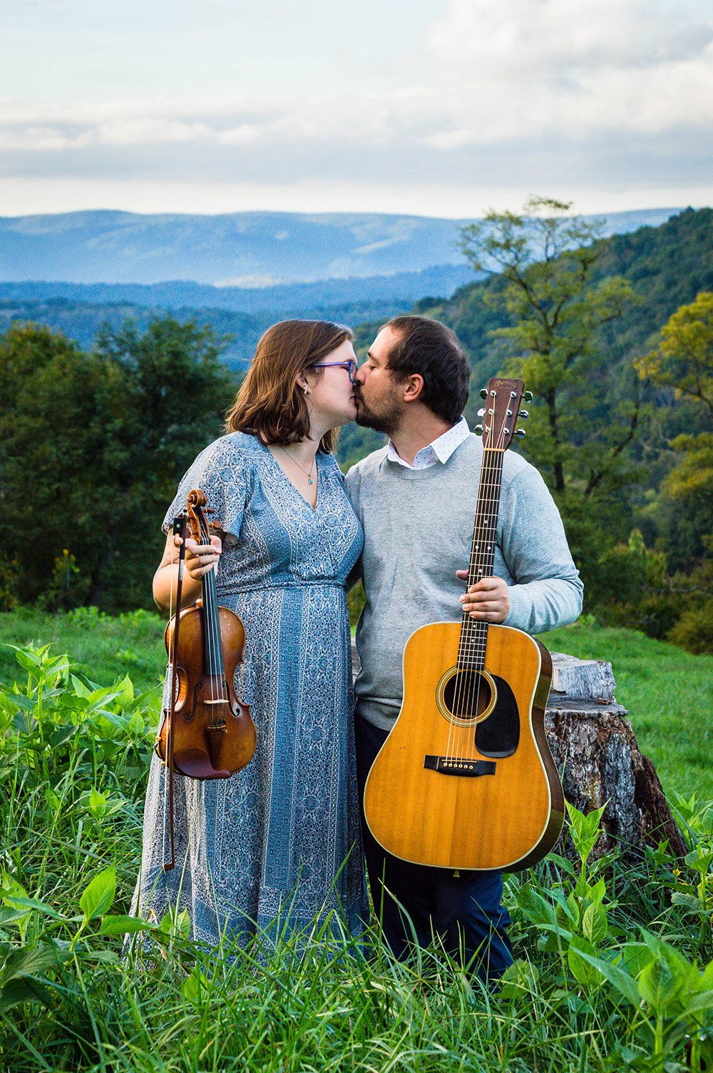 A couple holds a violin and guitar and go in for a kiss atop of a hill overlooking the Blue Ridge Mountains for a couple's adventure session in Blacksburg, Virginia.