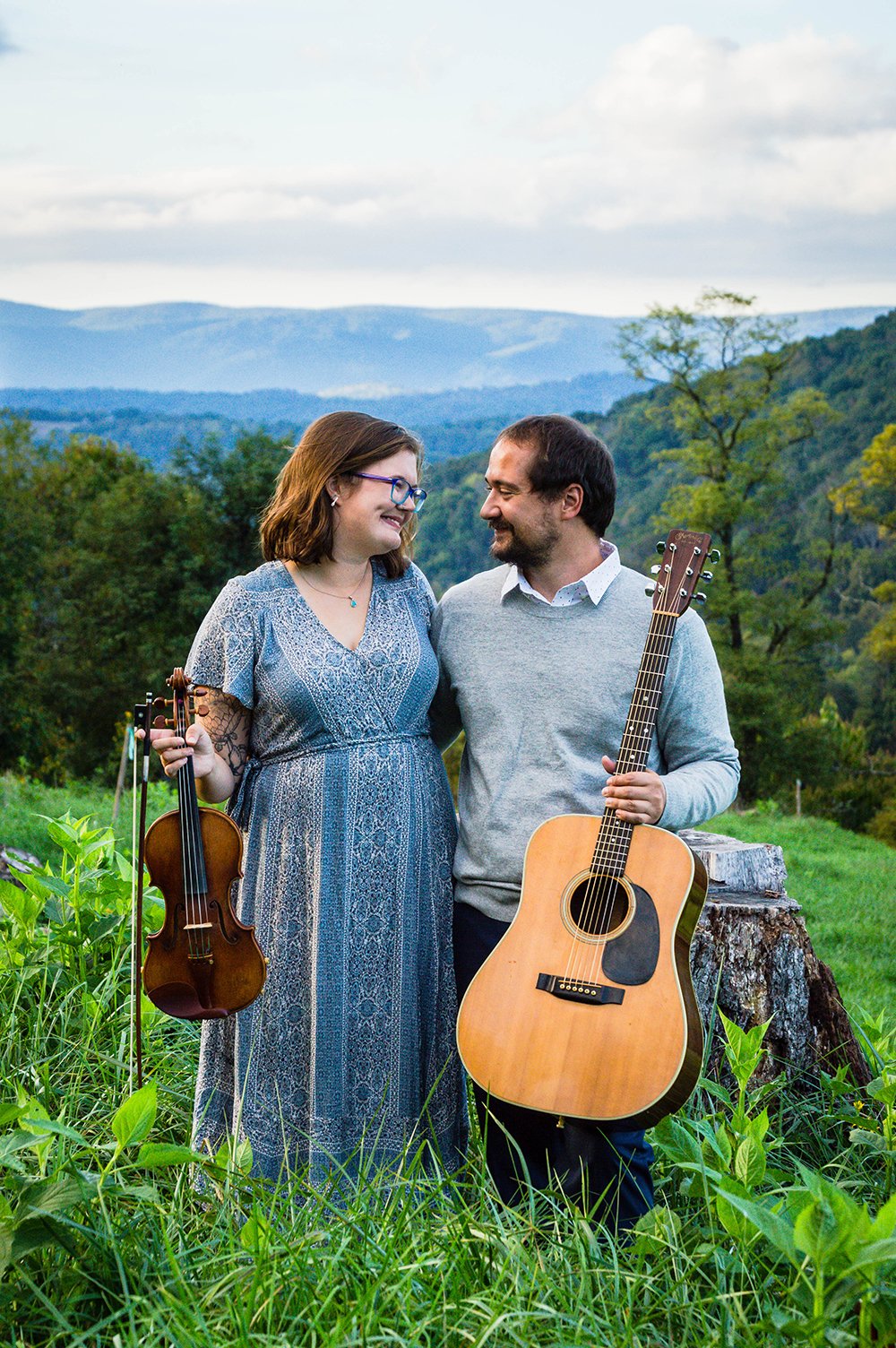 A couple holds a violin and guitar and smile lovingly at one another atop of a hill overlooking the Blue Ridge Mountains for a couple's adventure session in Blacksburg, Virginia.