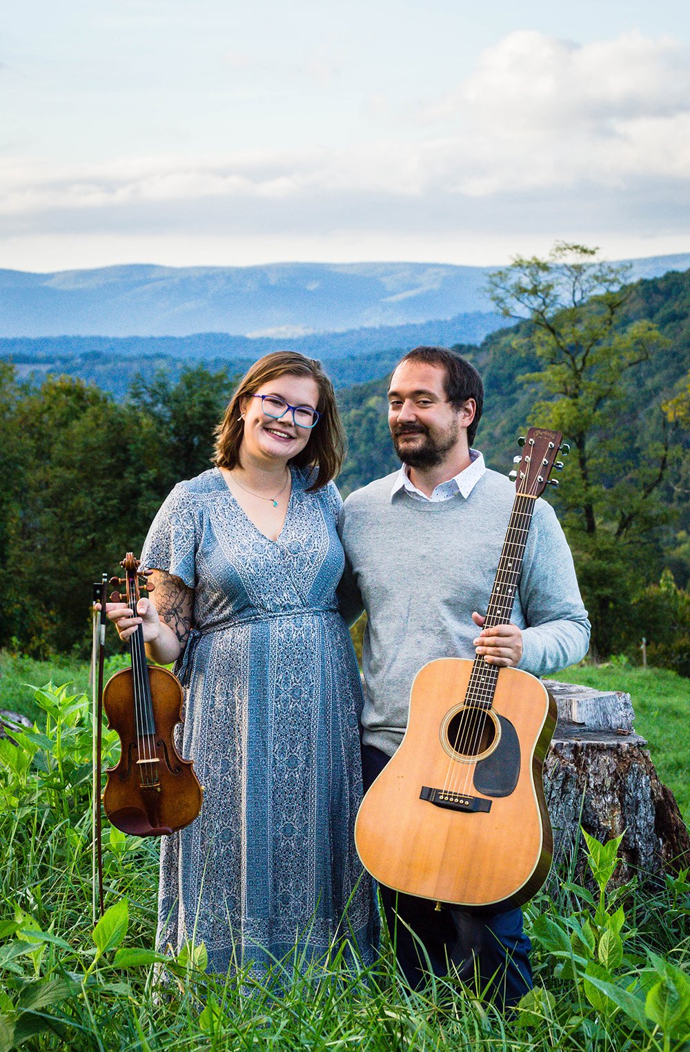 A couple holds a violin and guitar and smiles at the camera atop of a hill overlooking the Blue Ridge Mountains for a couple's adventure session in Blacksburg, Virginia.