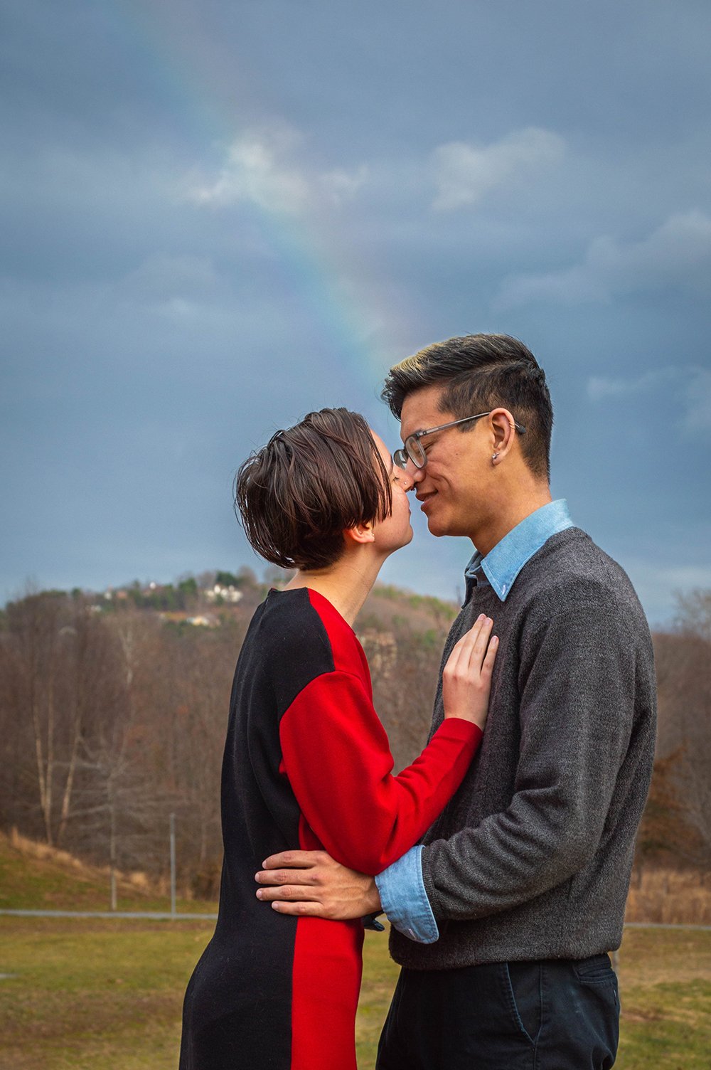 A couple goes in for a kiss as a rainbow appears in the sky.