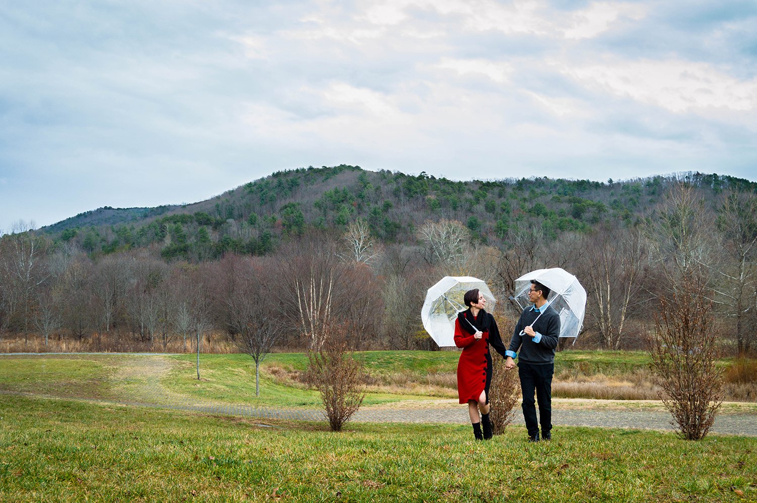 A couple walk hand in hand and look at one another and stick their tongues out as they walk up a hill with clear bubble umbrellas.