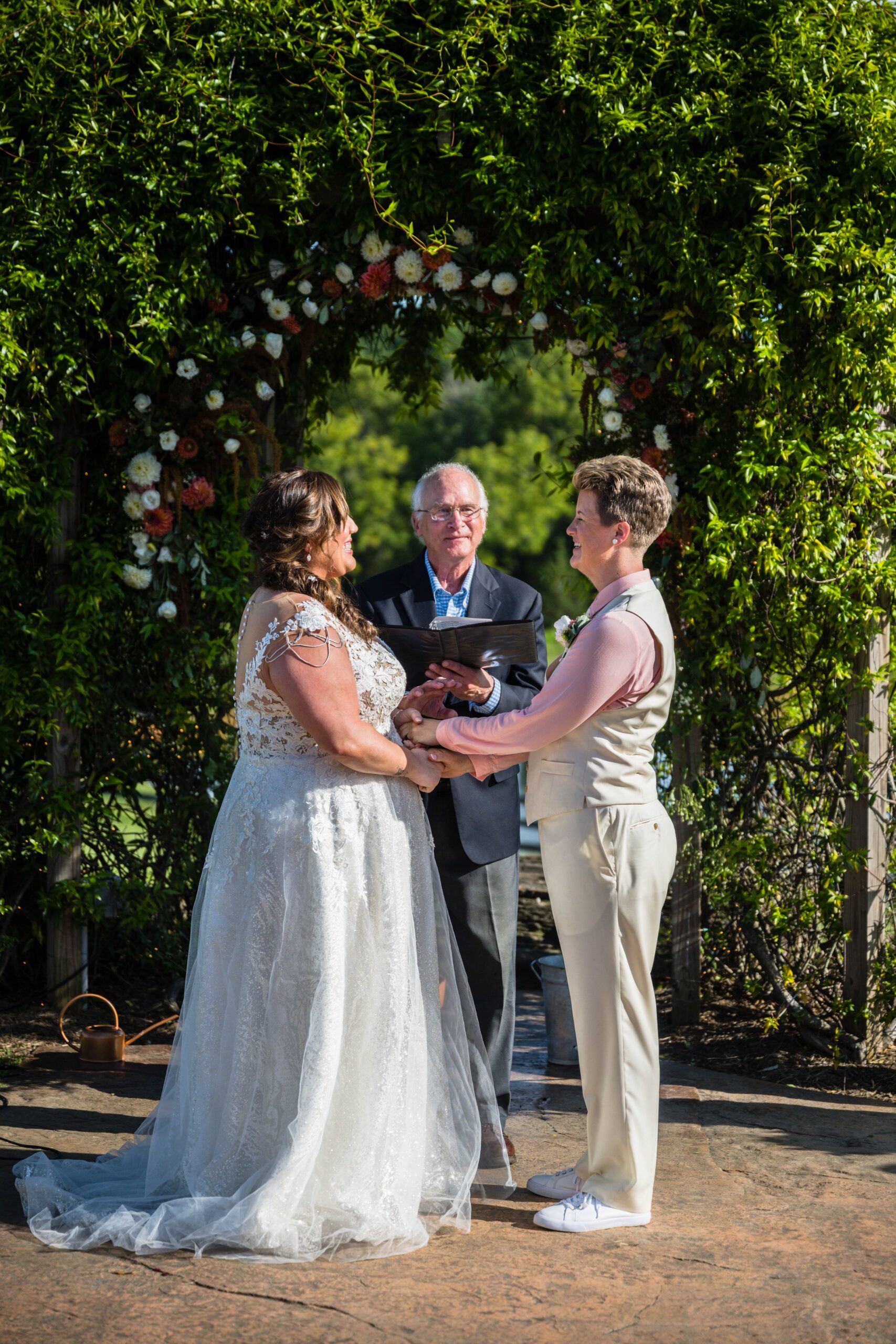 An LGBTQ+ couple stands at the altar holding hands and smiling at one another as their lgbtq officiant finishes the ceremony.