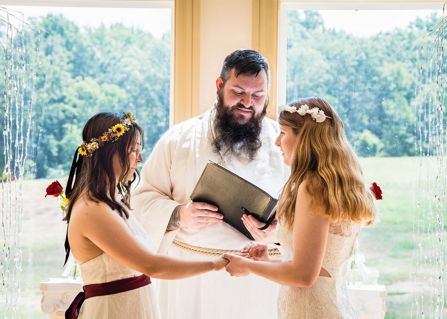 Two LGBTQ+ marriers hold hands in front of their LGBTQ officiant, who is reading something from his binder, while one of the marriers starts to place a ring on their partner's finger.