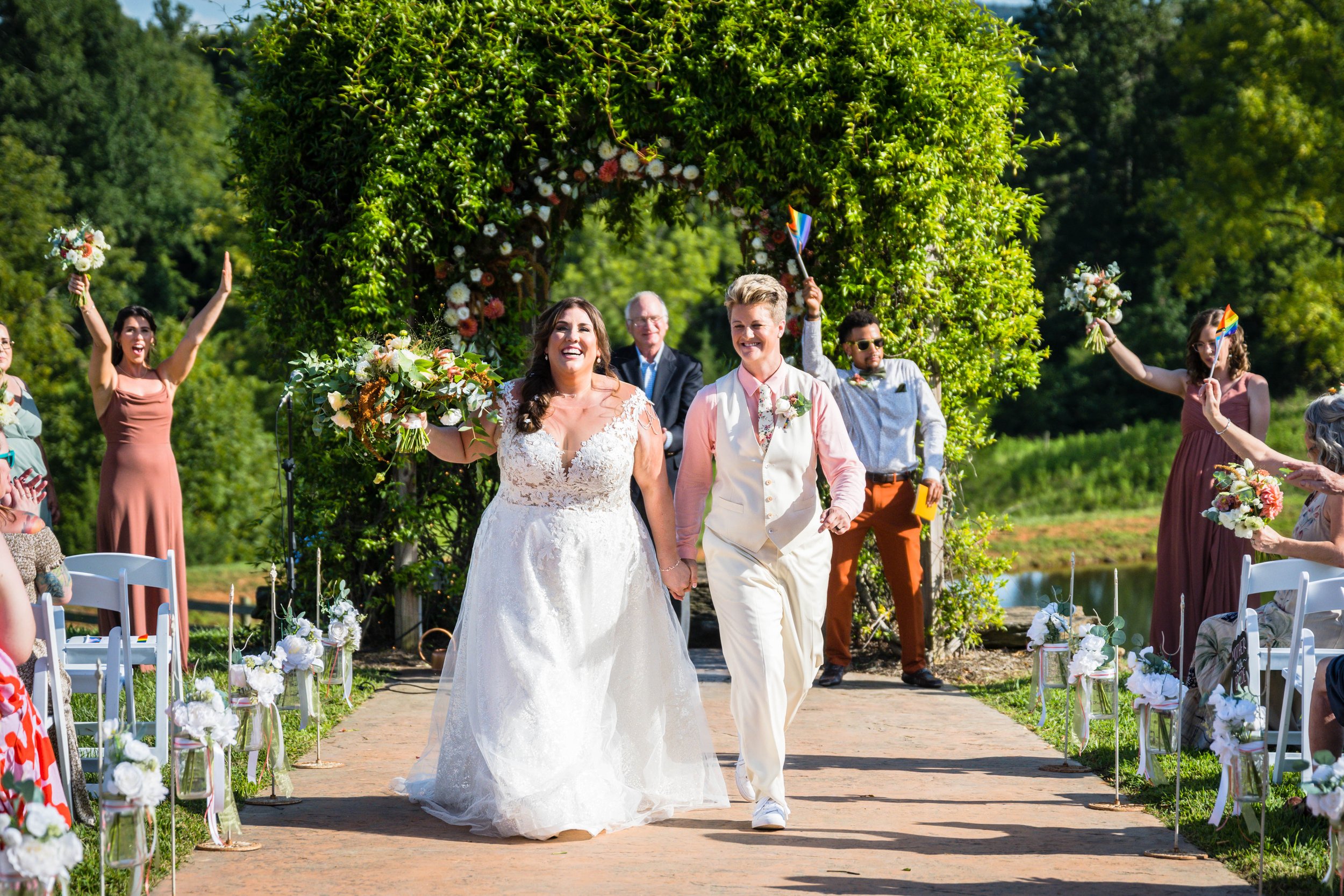 An lgbtq+ newlywed couple begin to walk away from the altar as their individual siblings cheer for both of them and lgbtq officiant smiles.