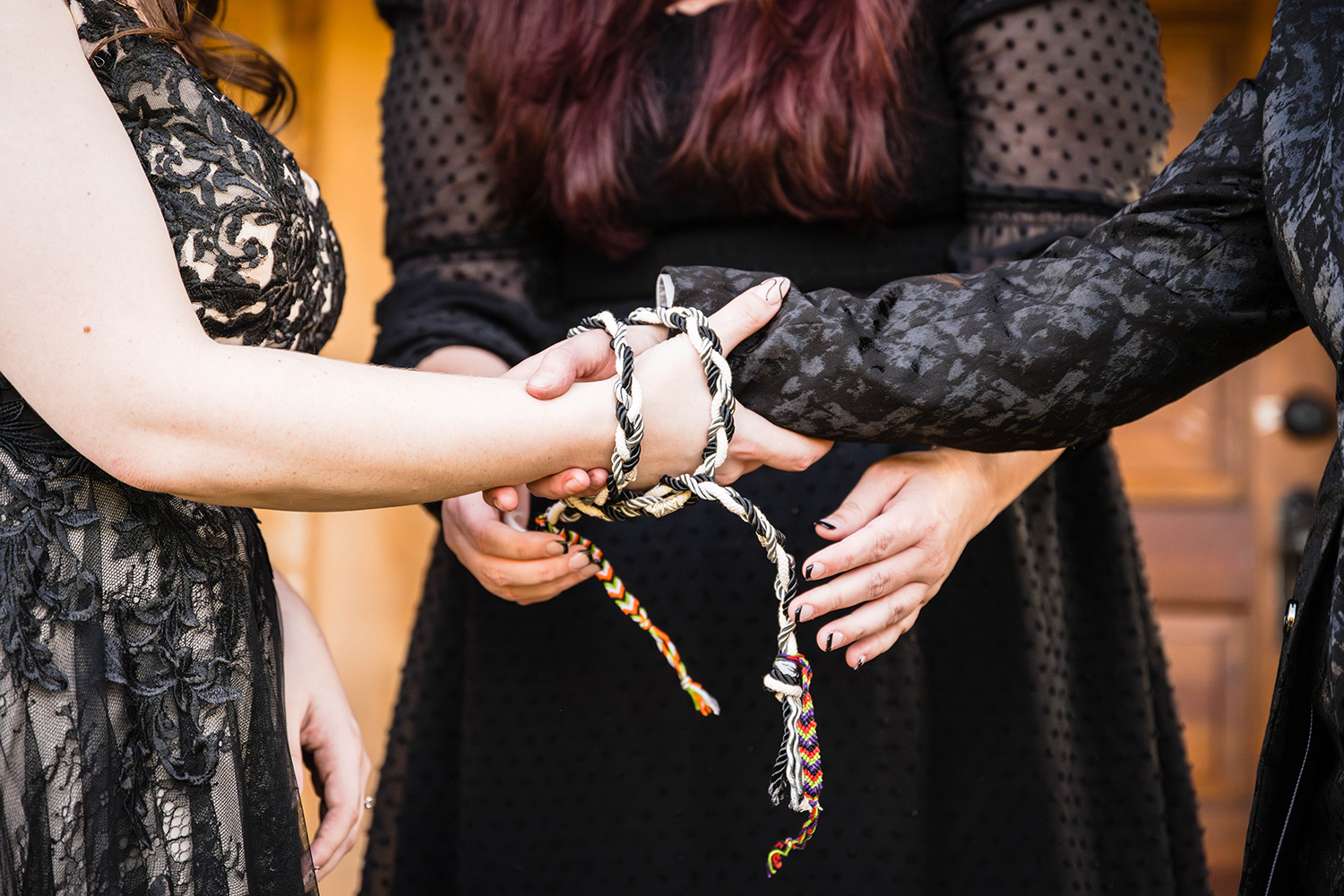 A queer friendly and affirming wedding officiant ties a braided rope around a couple's hands on their elopement day to perform a handfasting ceremony.