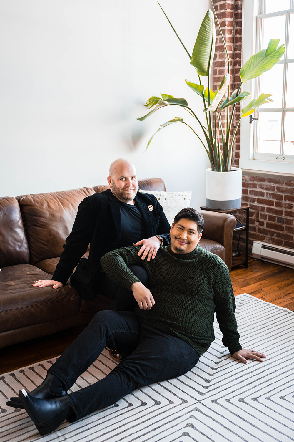 A gay couple sit inside of their living room and pose for a photo and grin towards the camera.