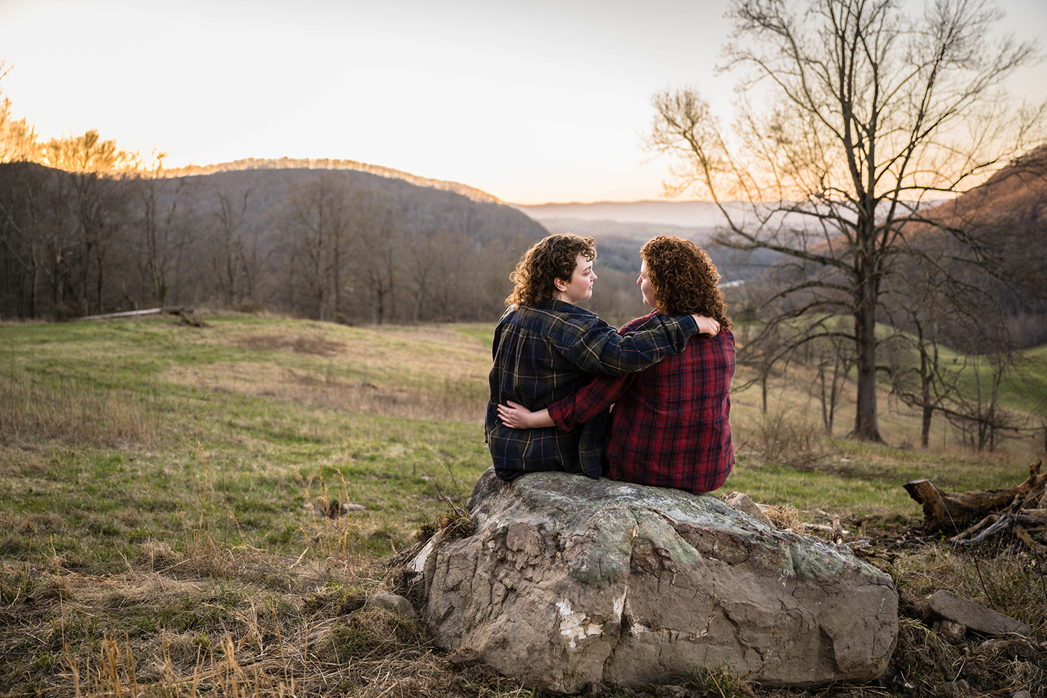 A lesbian couple sit on a large rock and look at one another with the sun setting quickly against the Blue Ridge Mountains in the background.