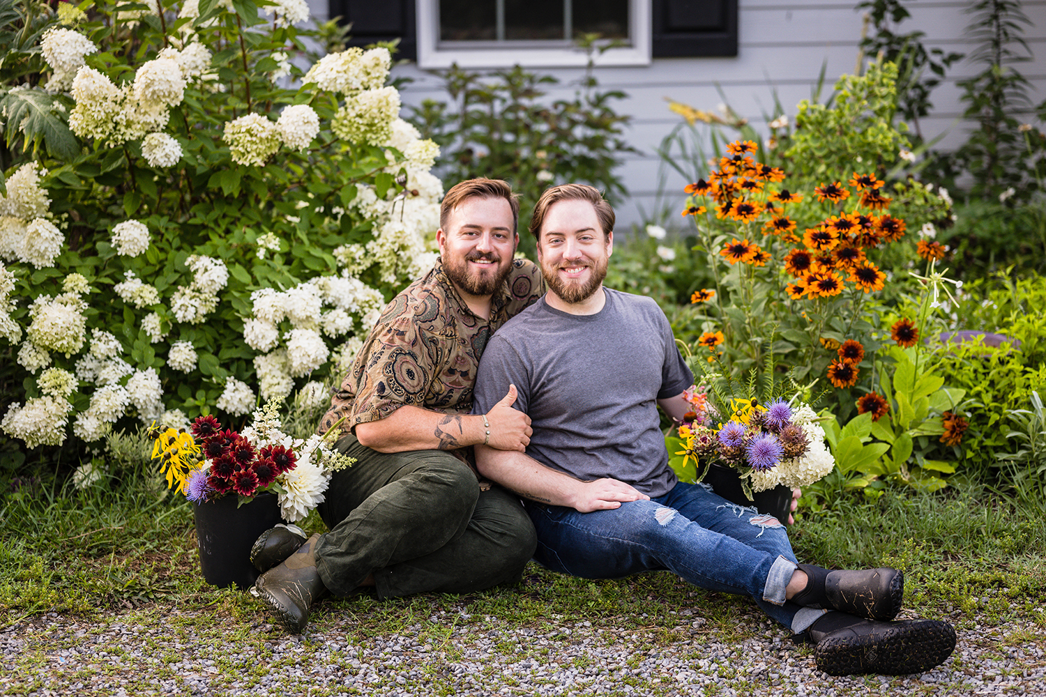 A gay couple sits outside of their home with buckets of flowers on either side of them. They sit closely to one another and smile towards the camera.