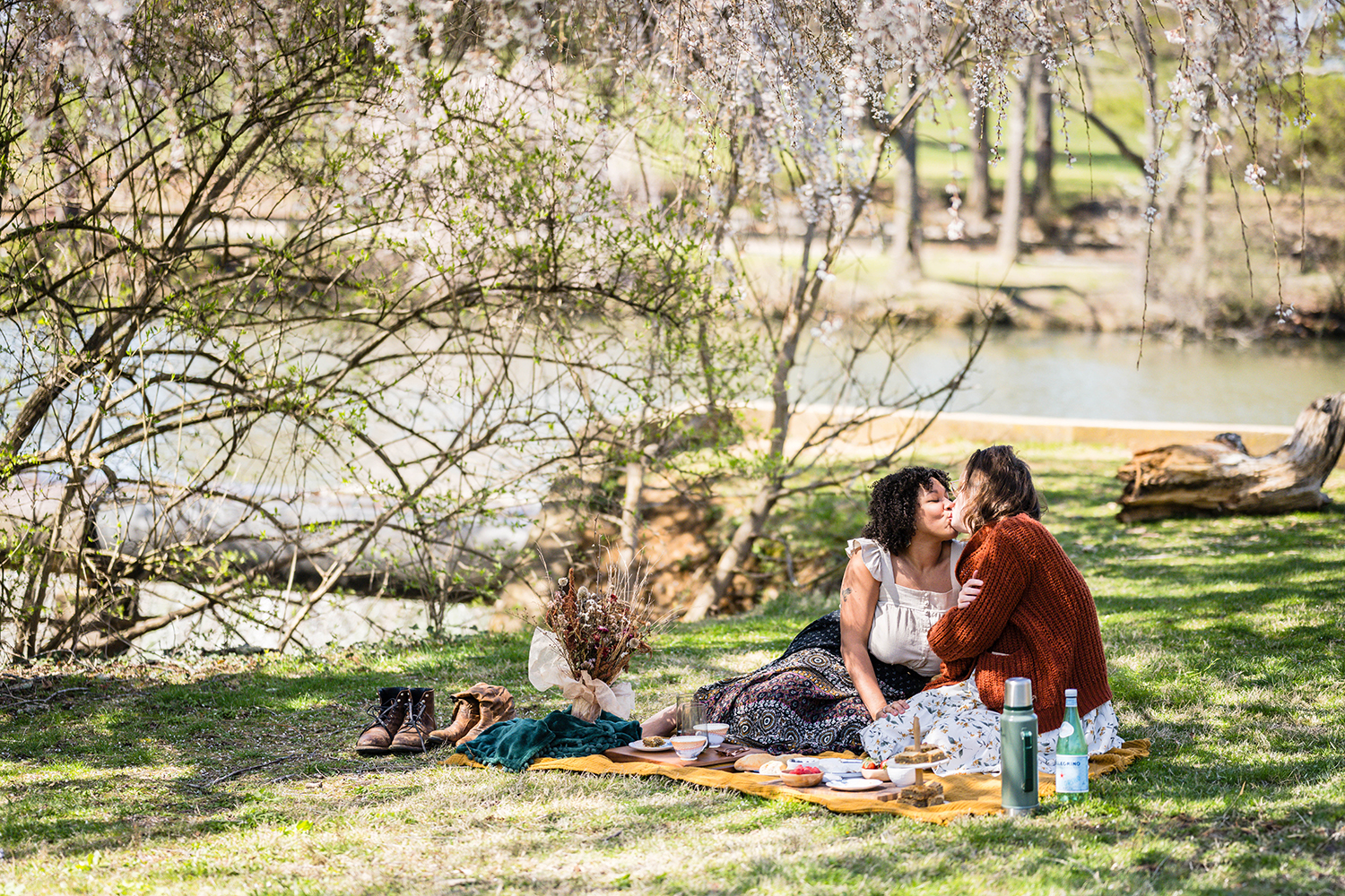 An LGBTQ+ couple sits under a few trees and has a picnic at Duck Pond in Blacksburg, Virginia. The couple lean in for a kiss.