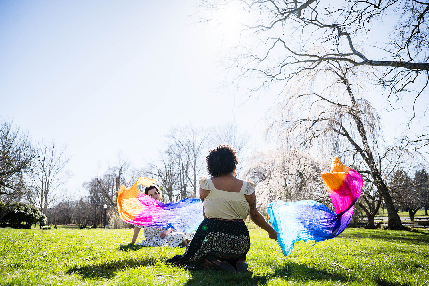 A trans marrier sits in the grass as her partner performs a fan dance on their elopement day at Duck Pond in Blacksburg, Virginia.