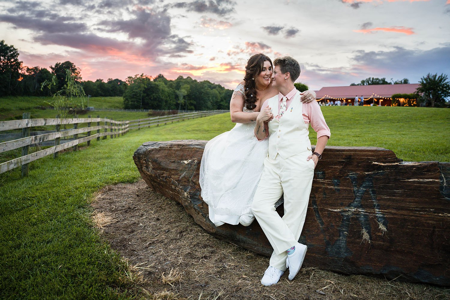 One lgbtq+ marrier sits atop a rock at the Pavilion at Black Water Junction while the other stands in front for a photo during their wedding day at sunset.