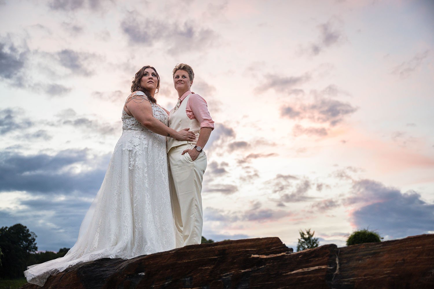 LGBTQ+ marriers stand atop a large rock at the Pavilion at Black Water Junction and hold onto one another and look away from the camera with a cloudy blue, purple, and pink sunset behind them.