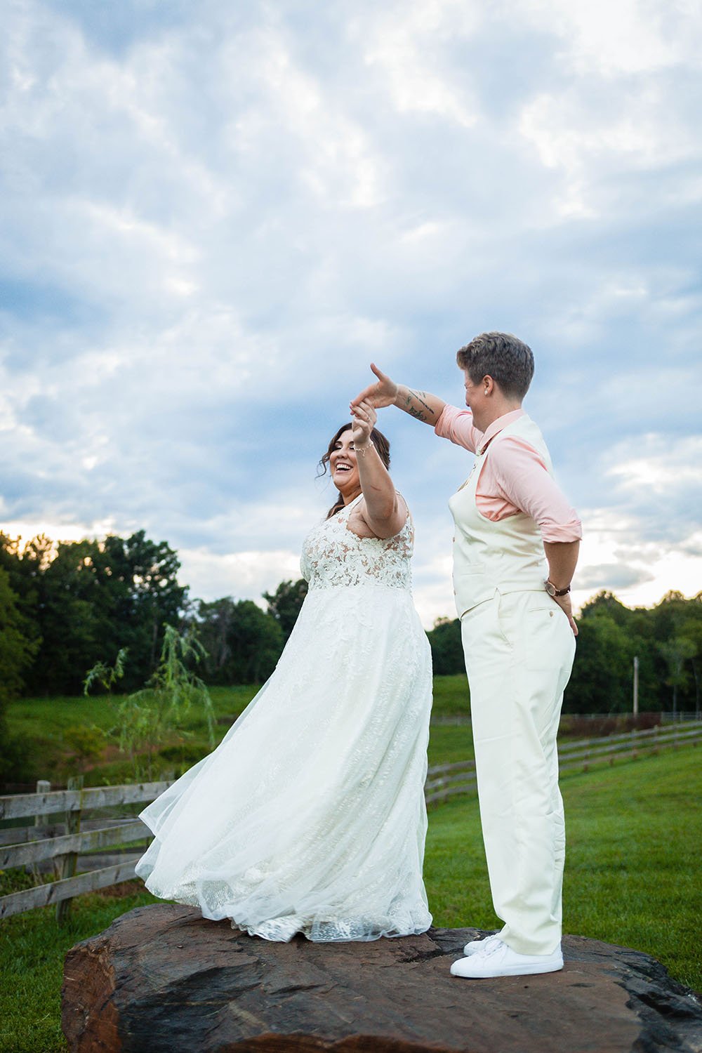 An lgbtq+ wedding couple dance on top of a large rock at the Pavilion at Black Water Junction under a cloudy, blue sky at sunset.