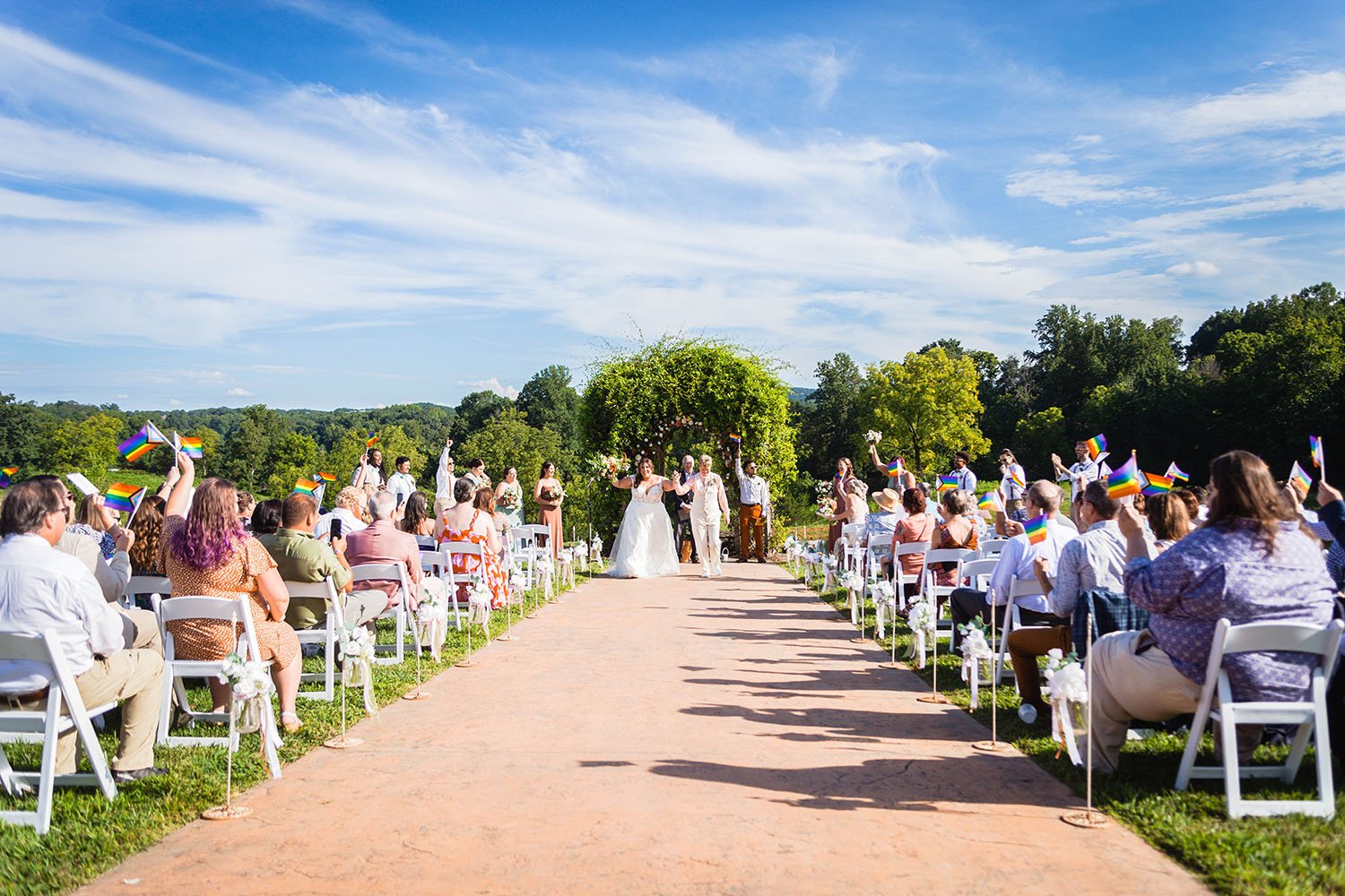 A wide shot of an lgbtq+ wedding ceremony at the Pavilion at Black Water Junction where the guests all wave rainbow flags.