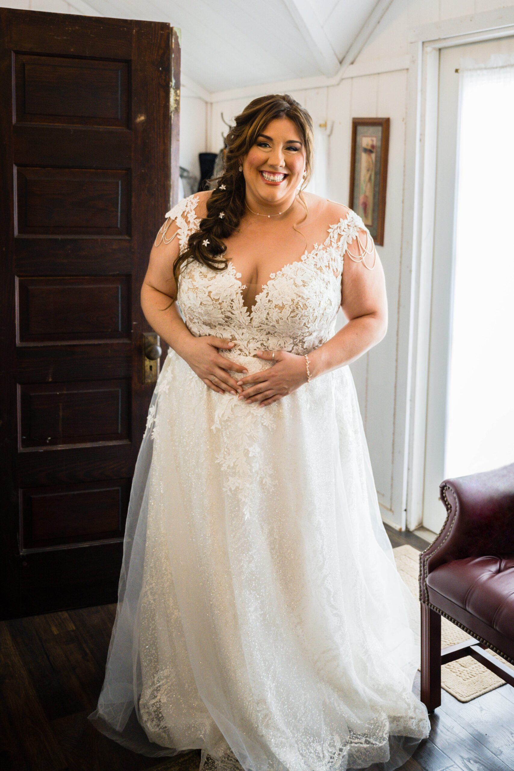 An lgbtq+ bride smiles wide as she reveals herself in her dress with hair and makeup done to her wedding party in a wedding suite.