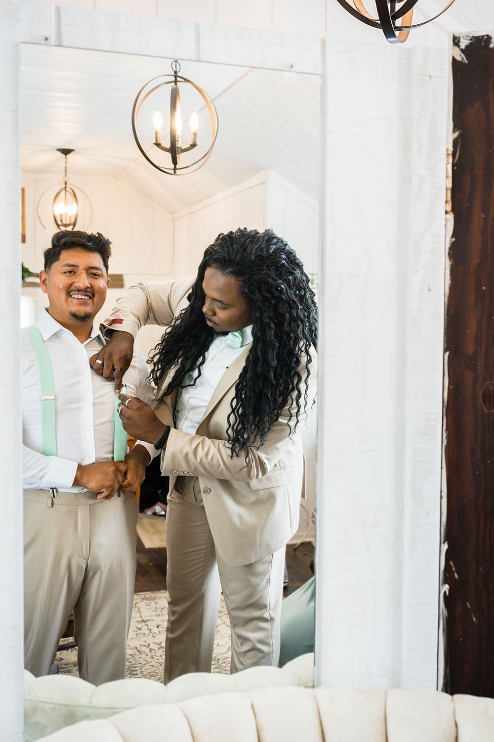 Two people face a mirror and adjust their suspenders and jackets during a wedding.