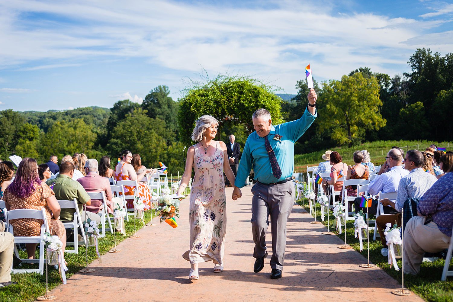 The mother and step-father of one of the marriers walk down the aisle holding hands while the step-father raises his fist holding a rainbow flag.