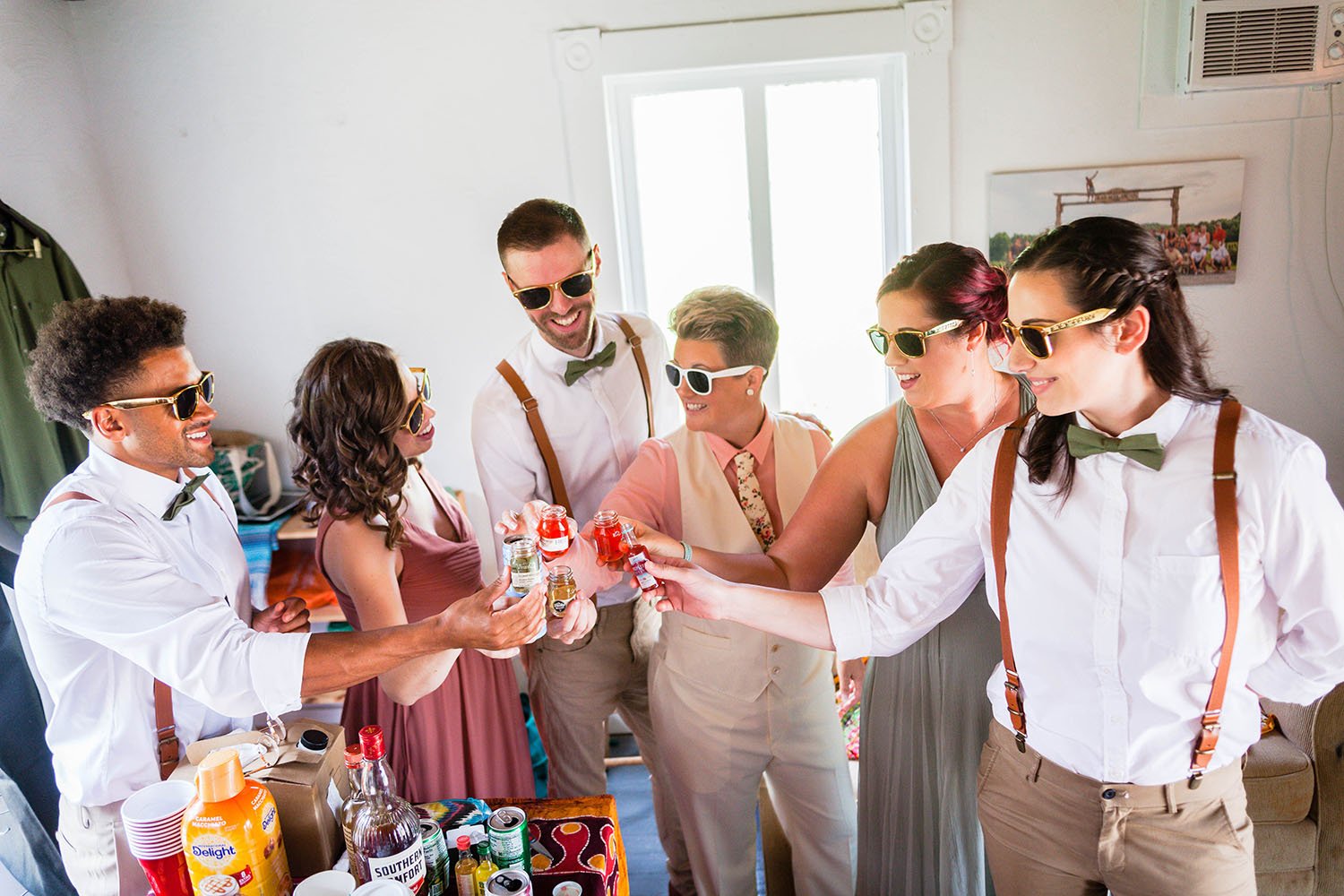 An lgbtq+ and mixed wedding party wear sunglasses inside of their wedding suite and hold out a drink to cheers the soon-to-be bride.