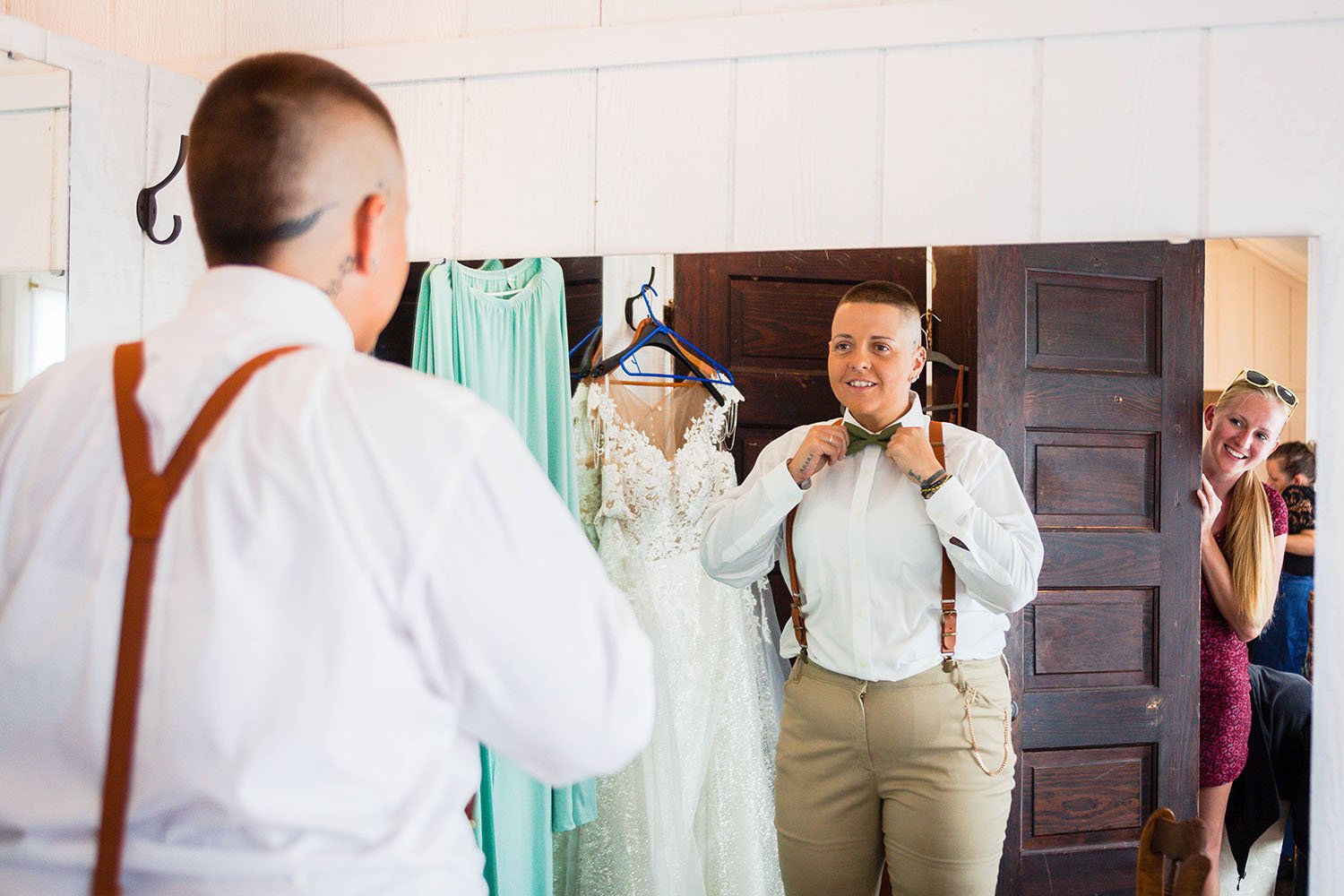 A person adjusts their bow tie in a wedding suite while their partner looks and smiles at them.