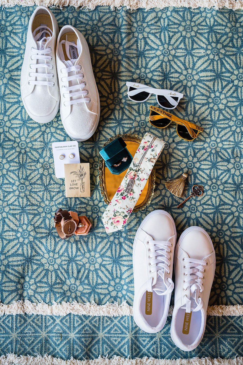 A birds eye view of a wedding details flatlay photograph featuring shoes, sunglasses, jewelry, and other accessories on a green patterned carpet.