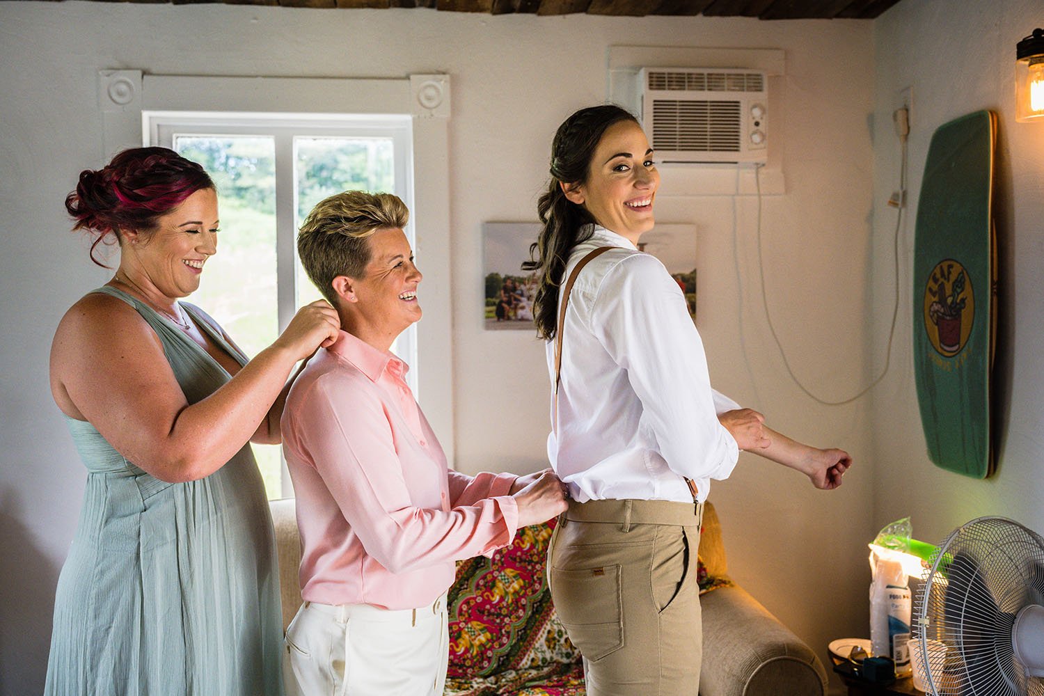 A group of three people stand in a line and help one another adjust their clothing as they get ready for a wedding.