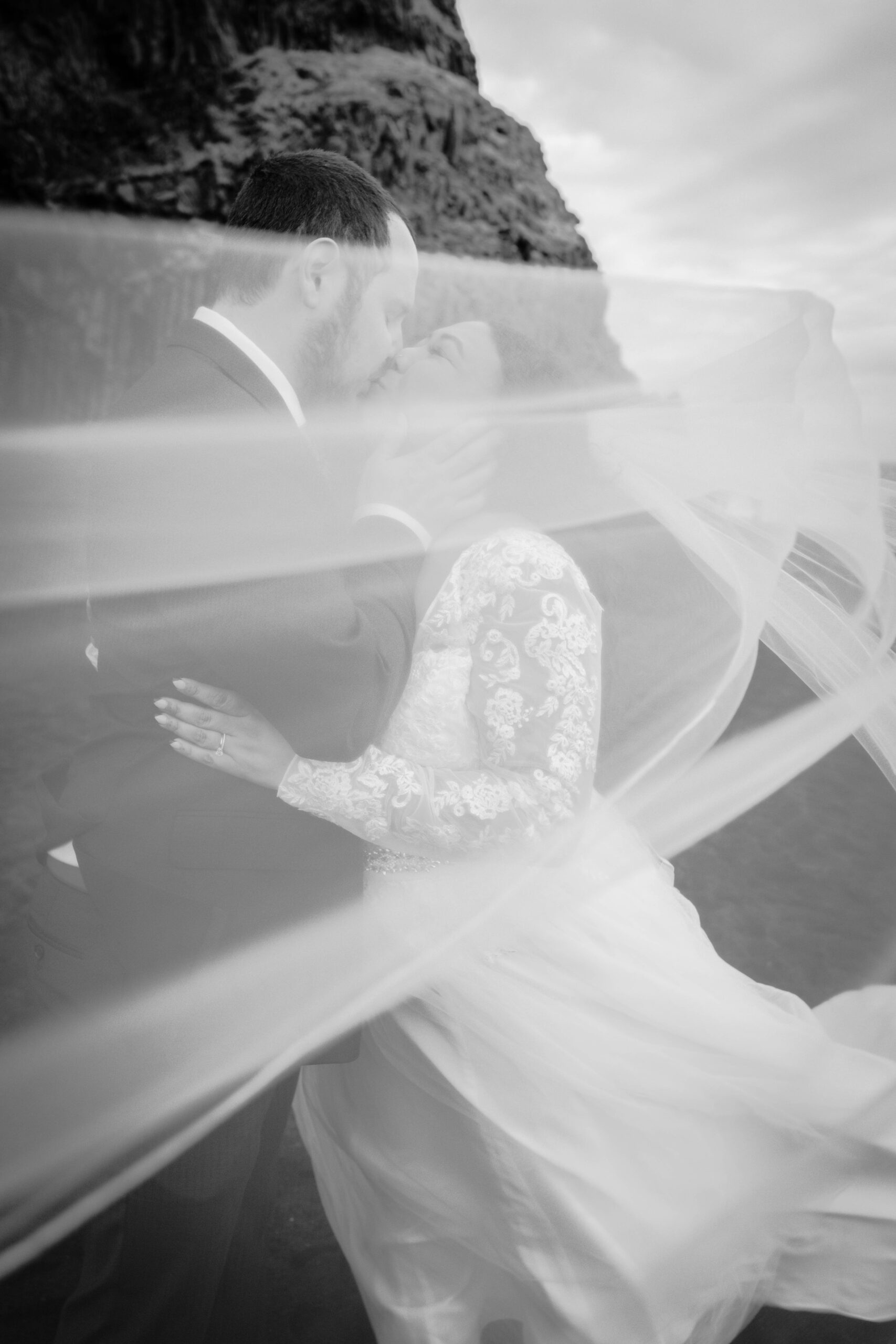 A newlywed couple kisses and embraces one another during their destination elopement at Reynisfjara Black Sands Beach in Iceland with the bride's veil whipping in front of the camera to create a dramatic moment.