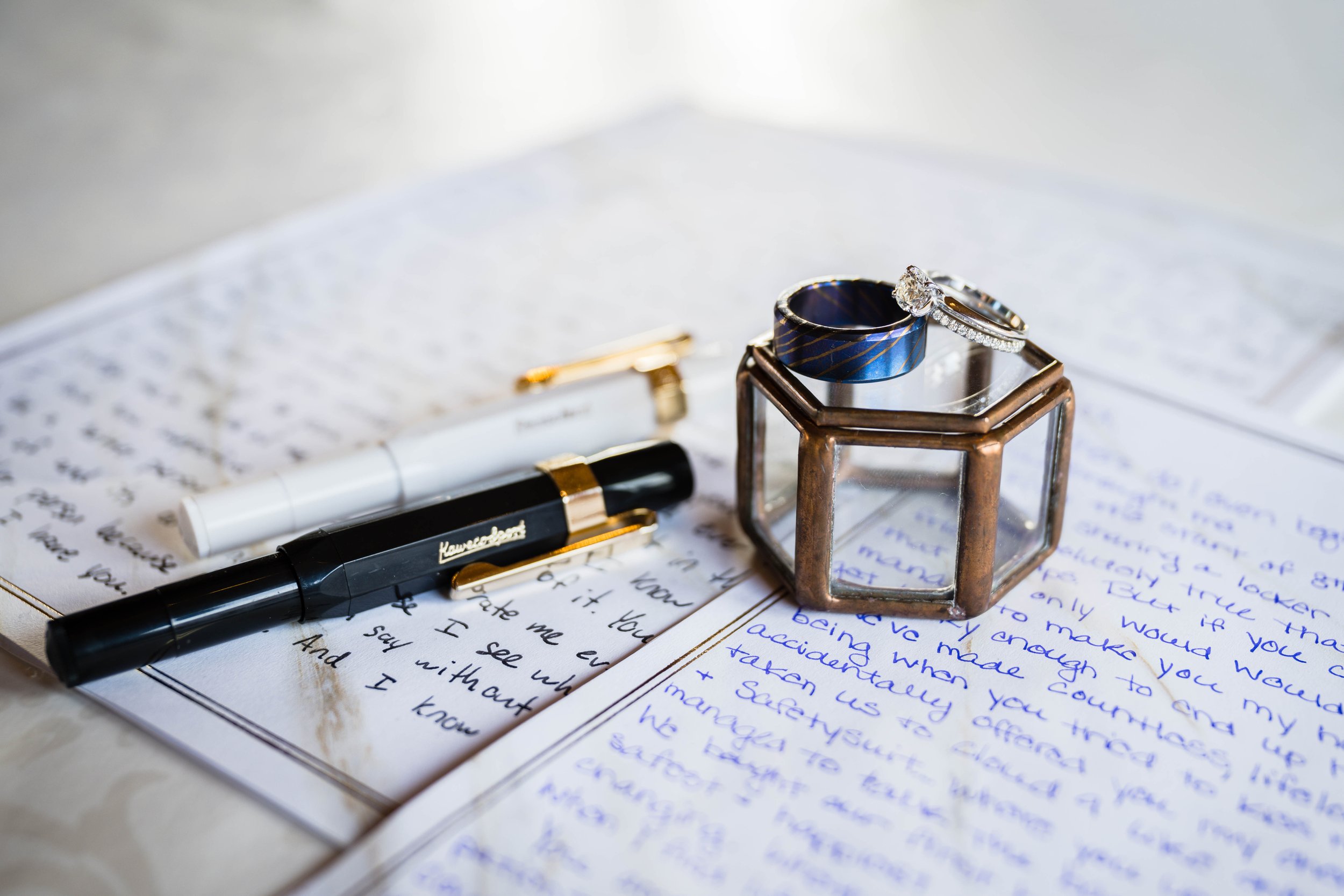 Written vows by a bride and groom lay on a table with white and black pens next to a clear ring box with two wedding rings propped on top.