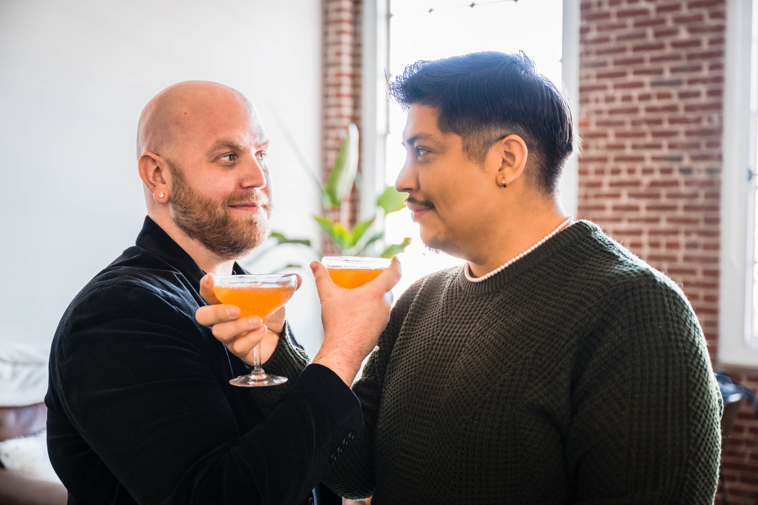 A gay couple intertwine their arms, each holding a cocktail in hand, and gaze at one another determinedly.