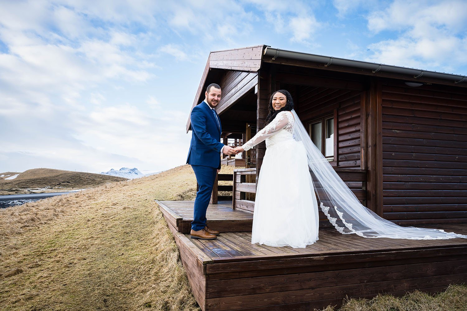 A wedding couple stands to the side of their cabin extending their hands towards one another and look over their shoulder and smile.