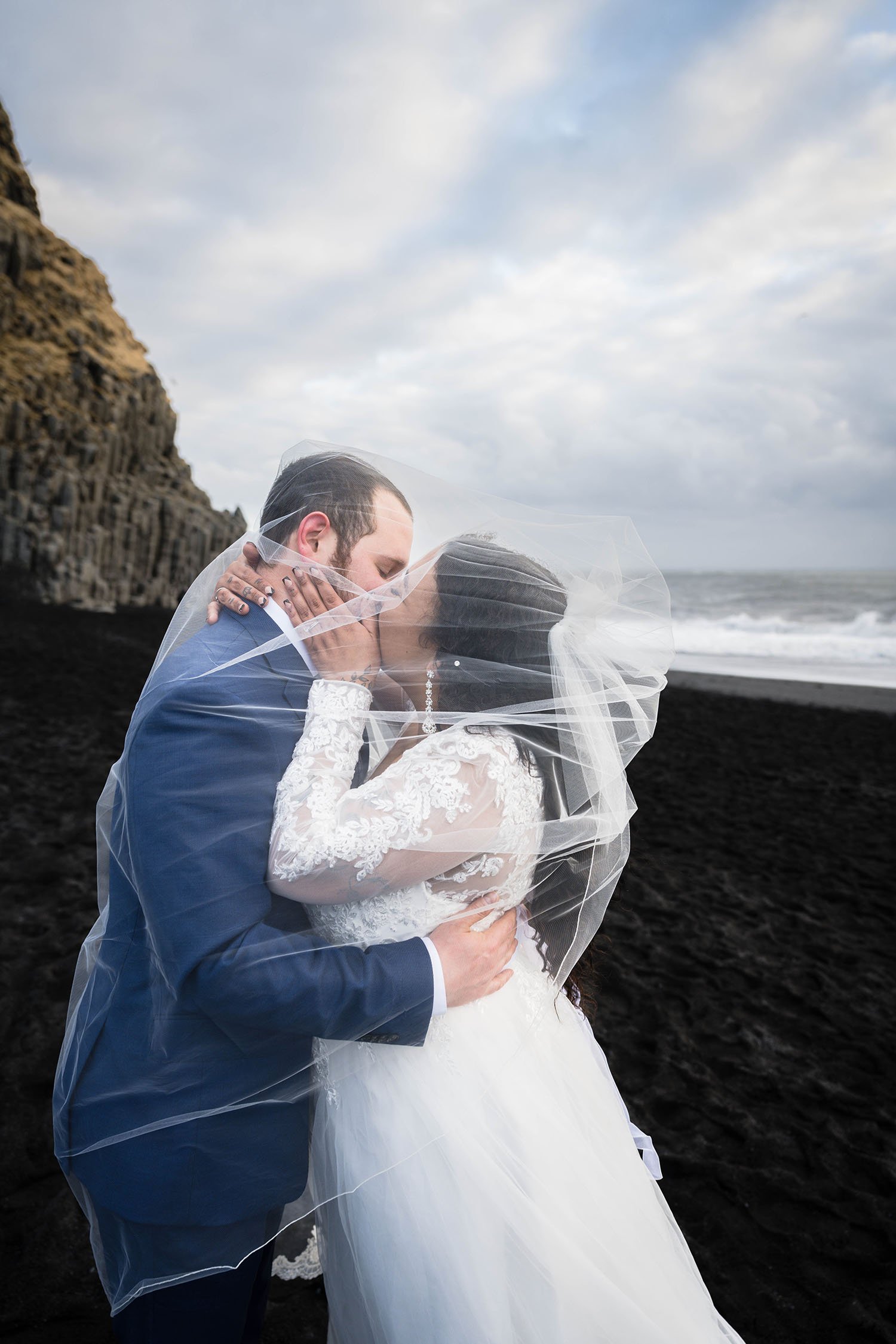 A couple eloping on Reynisfjara Beach in Iceland embrace for another kiss under a wedding veil.