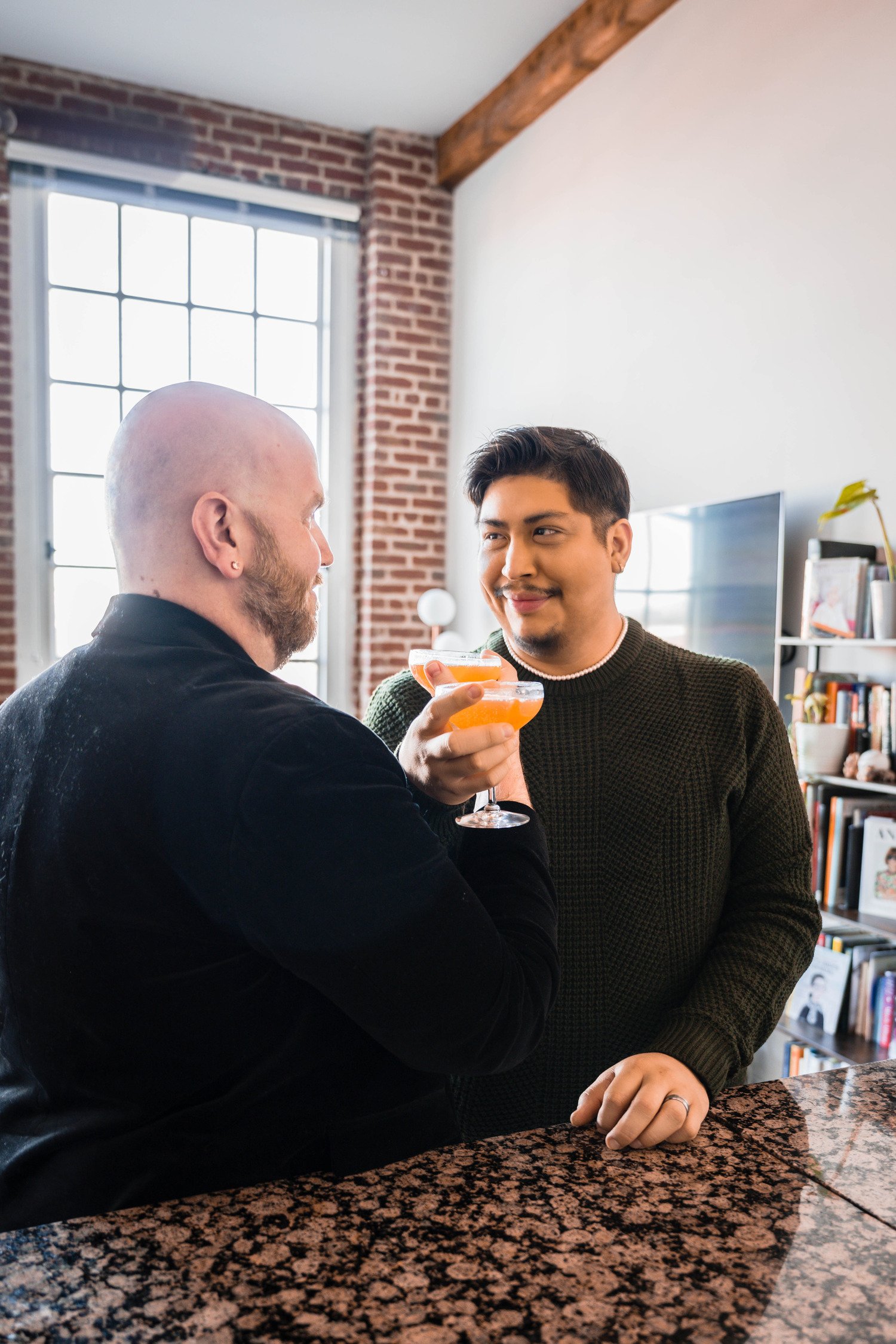 A queer couple make cocktail drinks and intertwine their arms as they go for a toast during their at-home photoshoot in Downtown Roanoke.