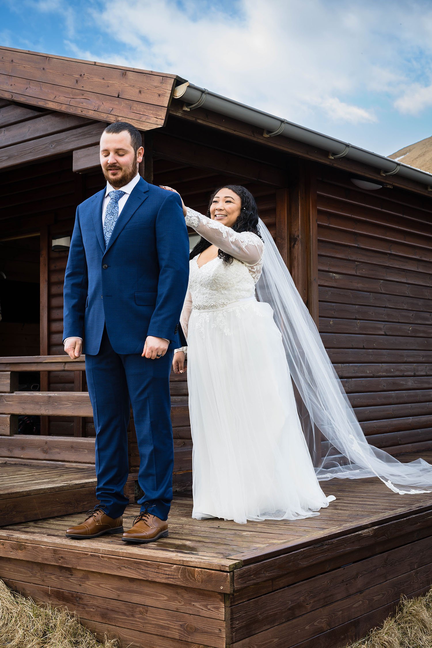 A woman in a wedding dress taps the shoulder of her partner wearing a suit outside of their cabin in Iceland.