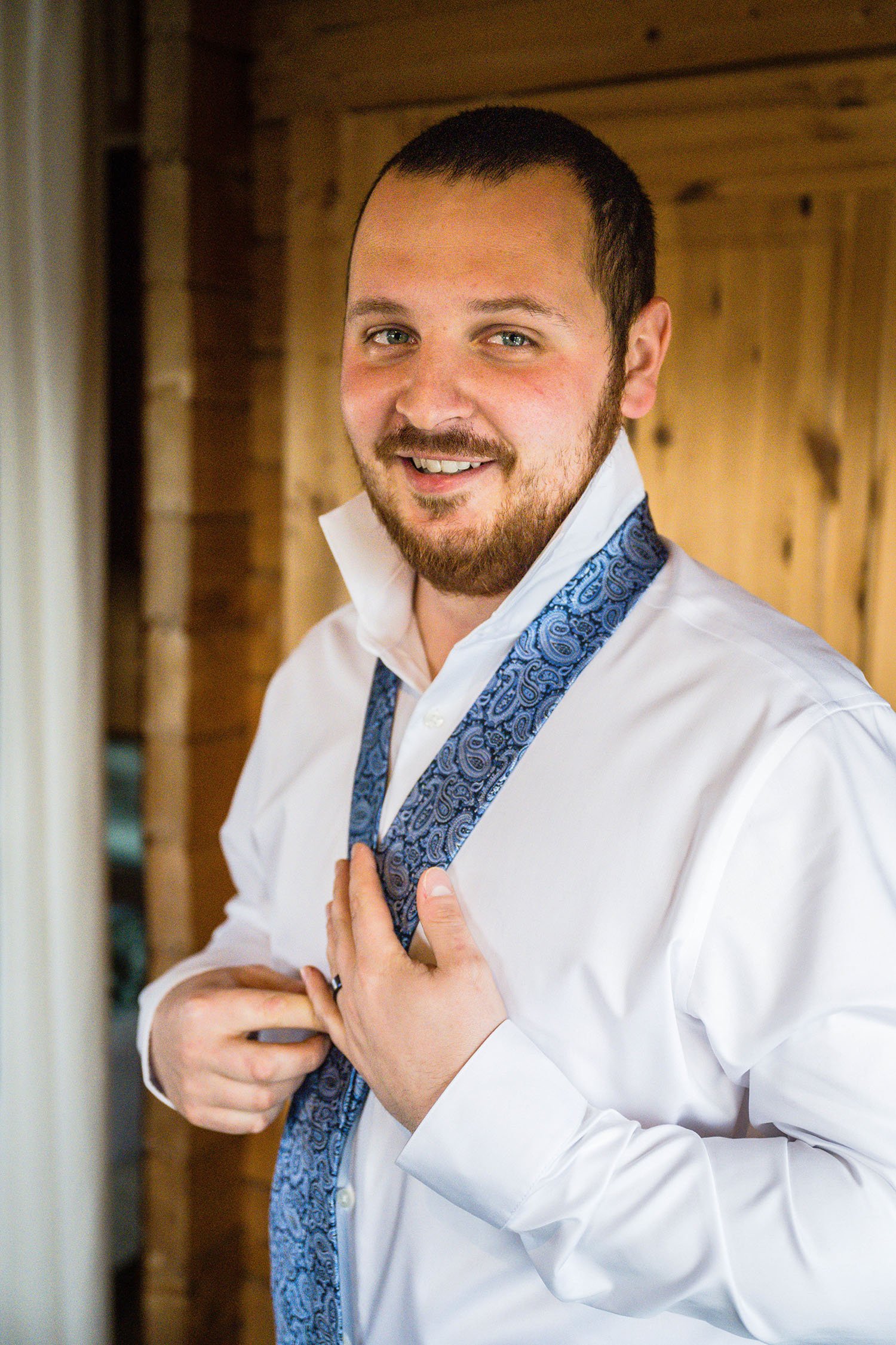 A man inside a wood cabin in Iceland ties a knot in his tie and smiles.