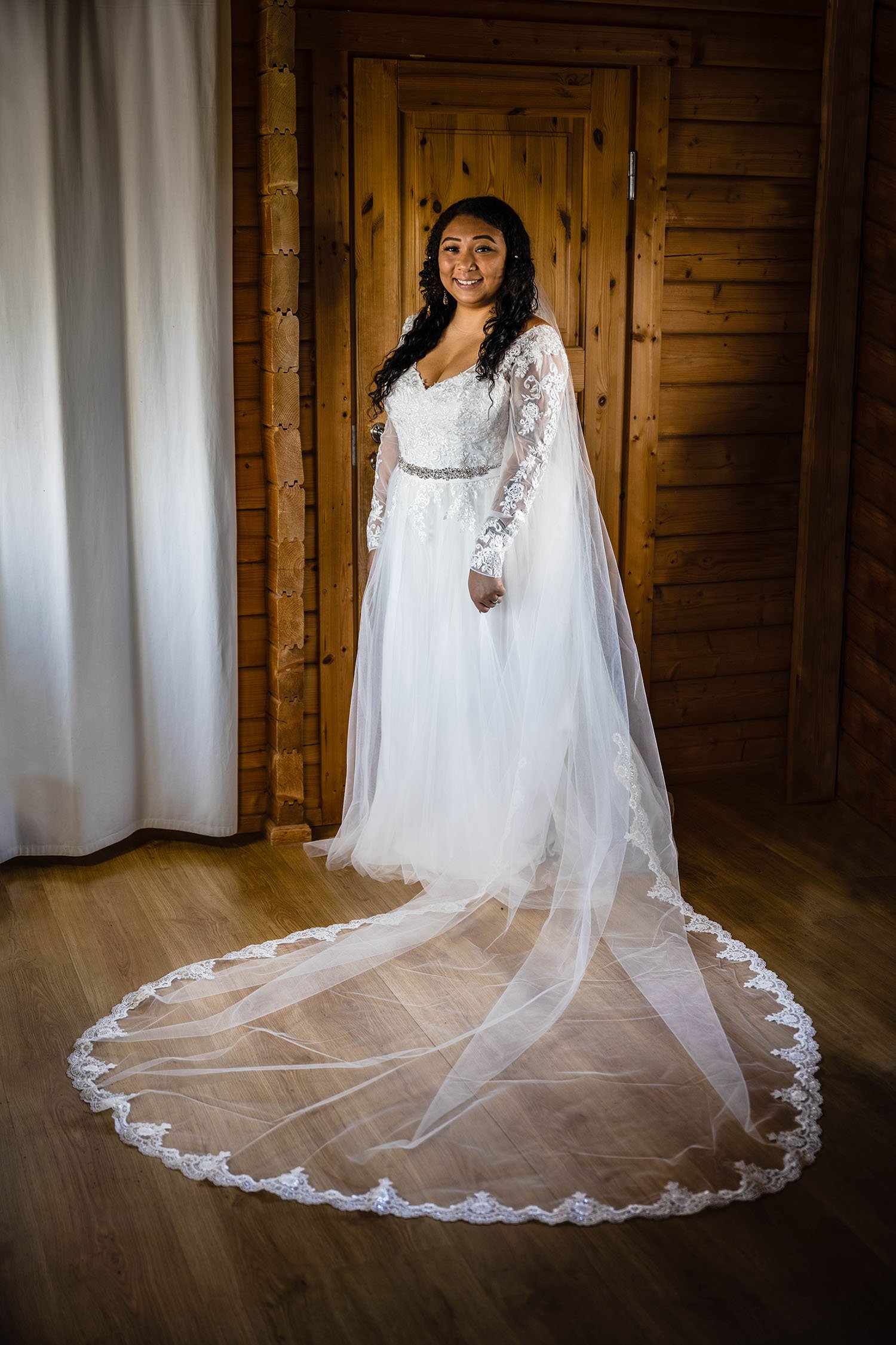 A woman wearing a wedding dress and long veil poses against a cabin door.