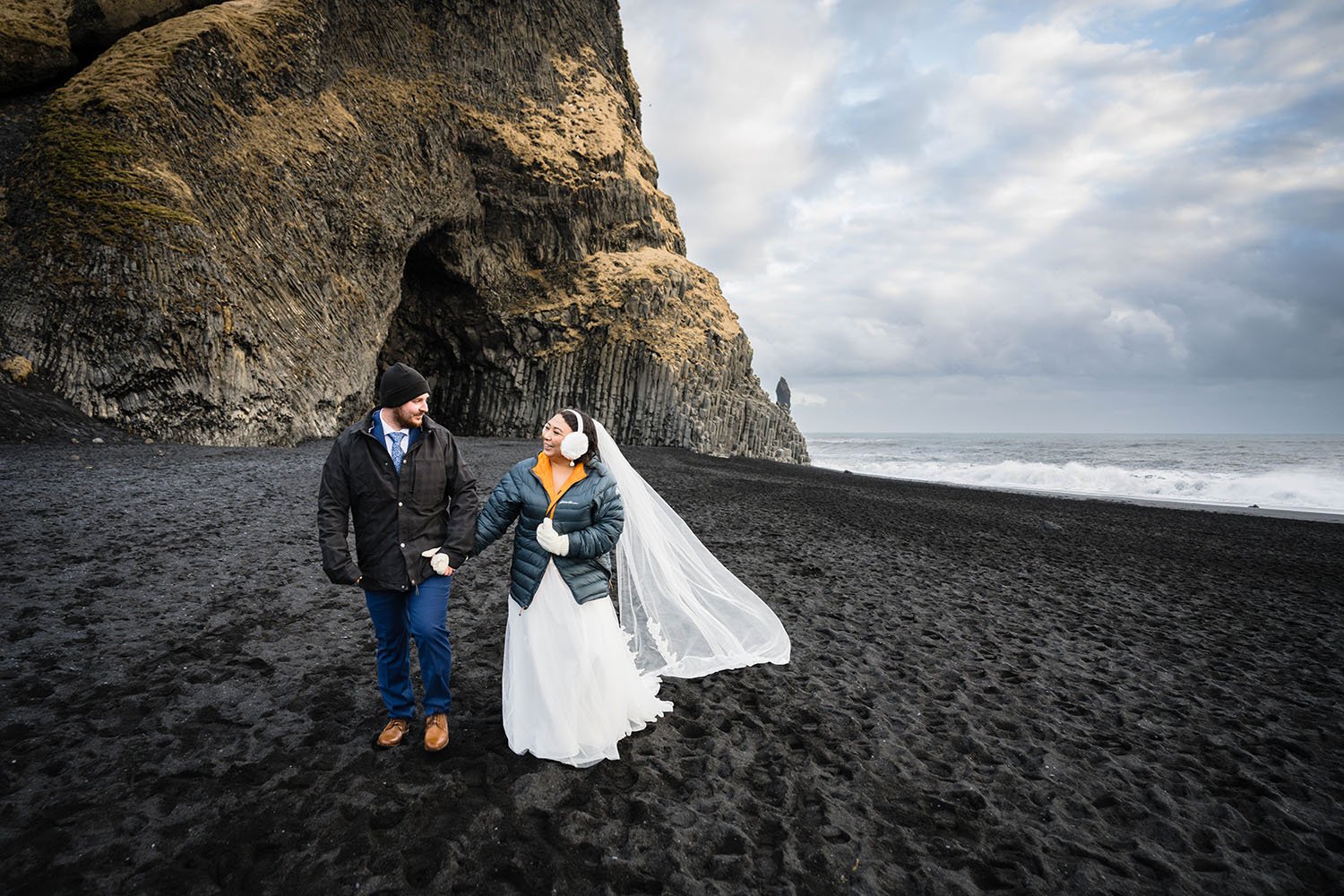 A couple eloping in Iceland looks at one another and walks hand in hand away from Hálsanefshellir Cave on Reynisfjara Beach in Iceland. 