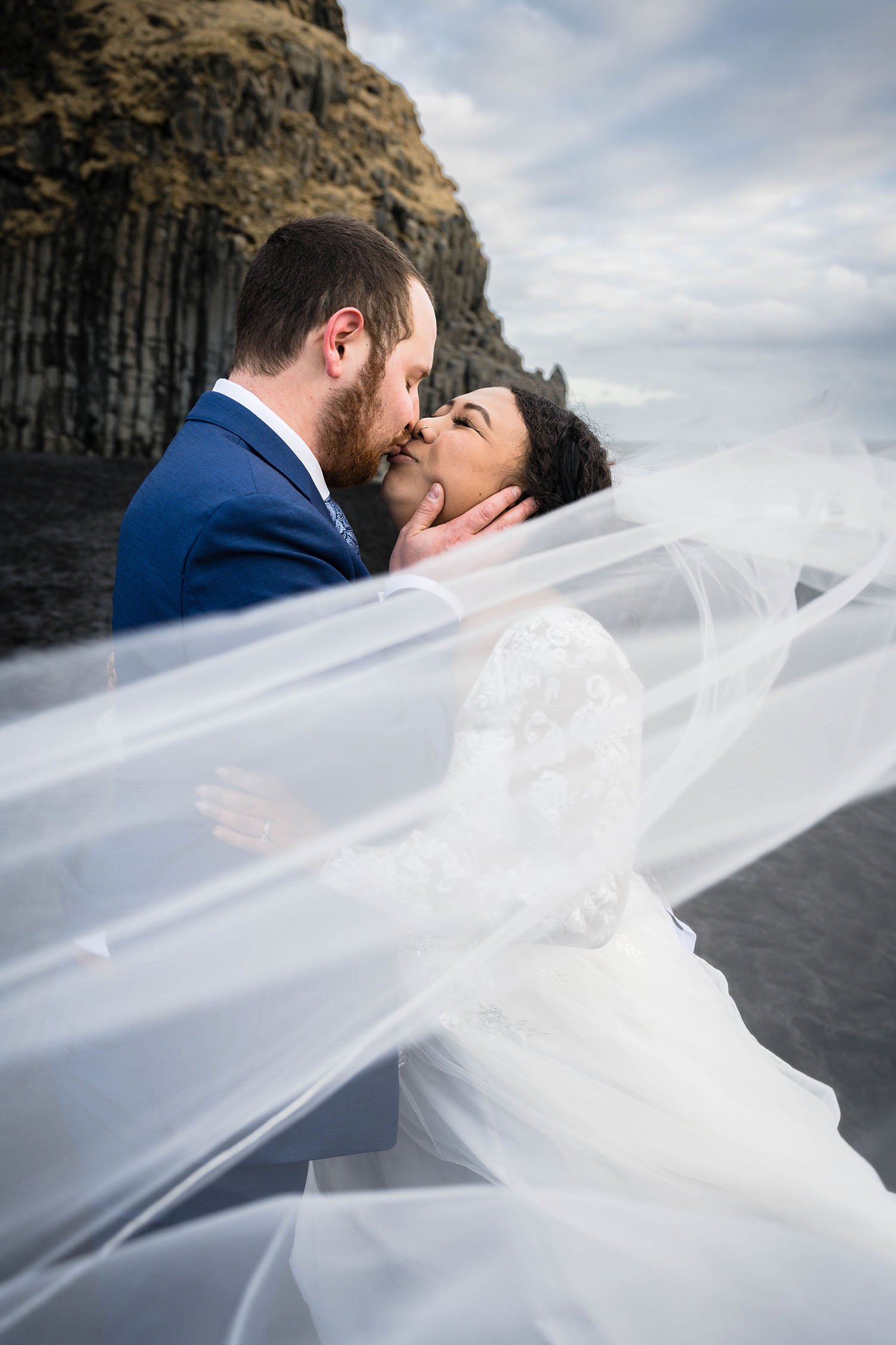 A couple eloping go in for a kiss with a wedding veil in the foreground on Reynisfjara Beach in Iceland.