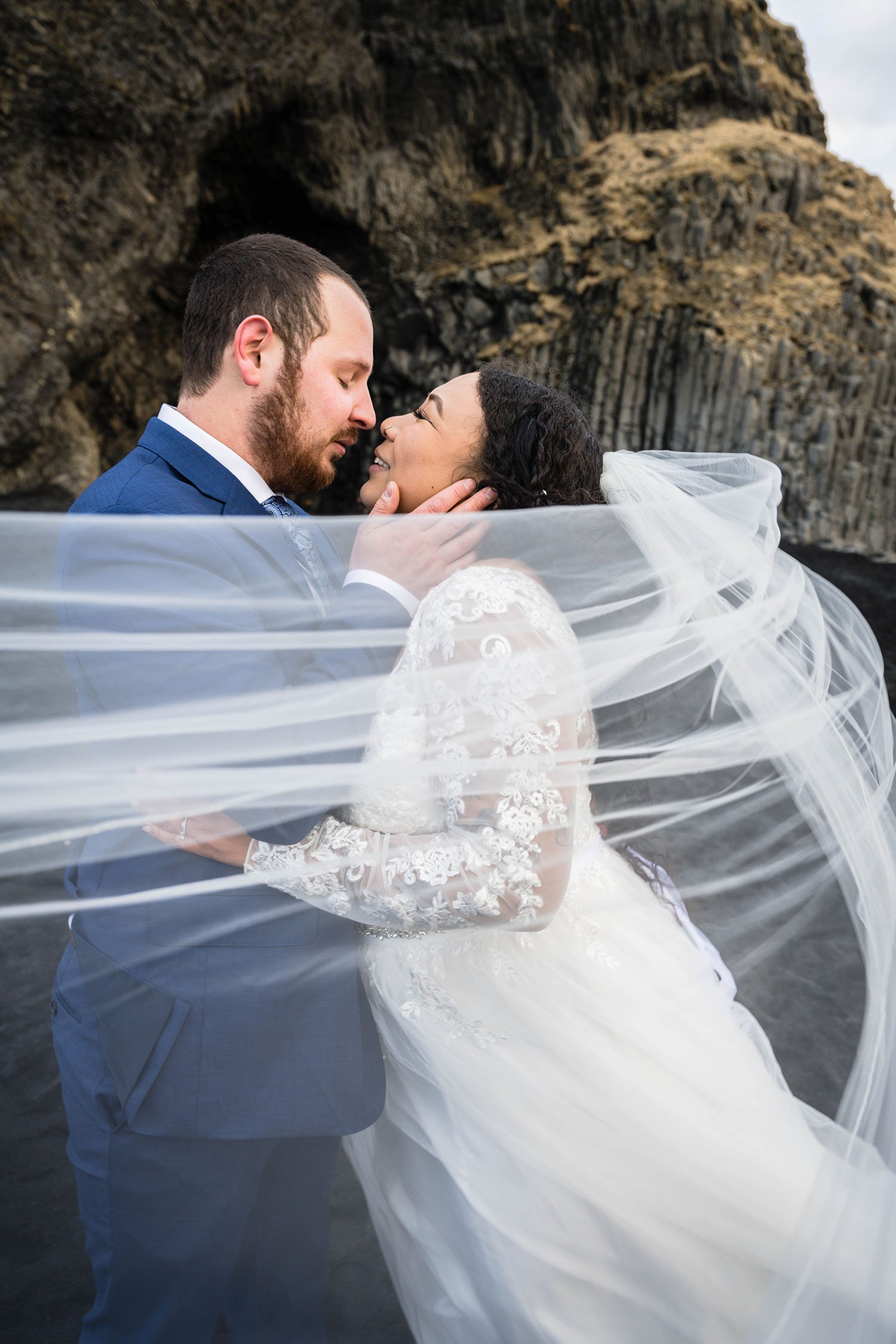 A couple eloping go in for a kiss with a wedding veil in the foreground on Reynisfjara Beach in Iceland.