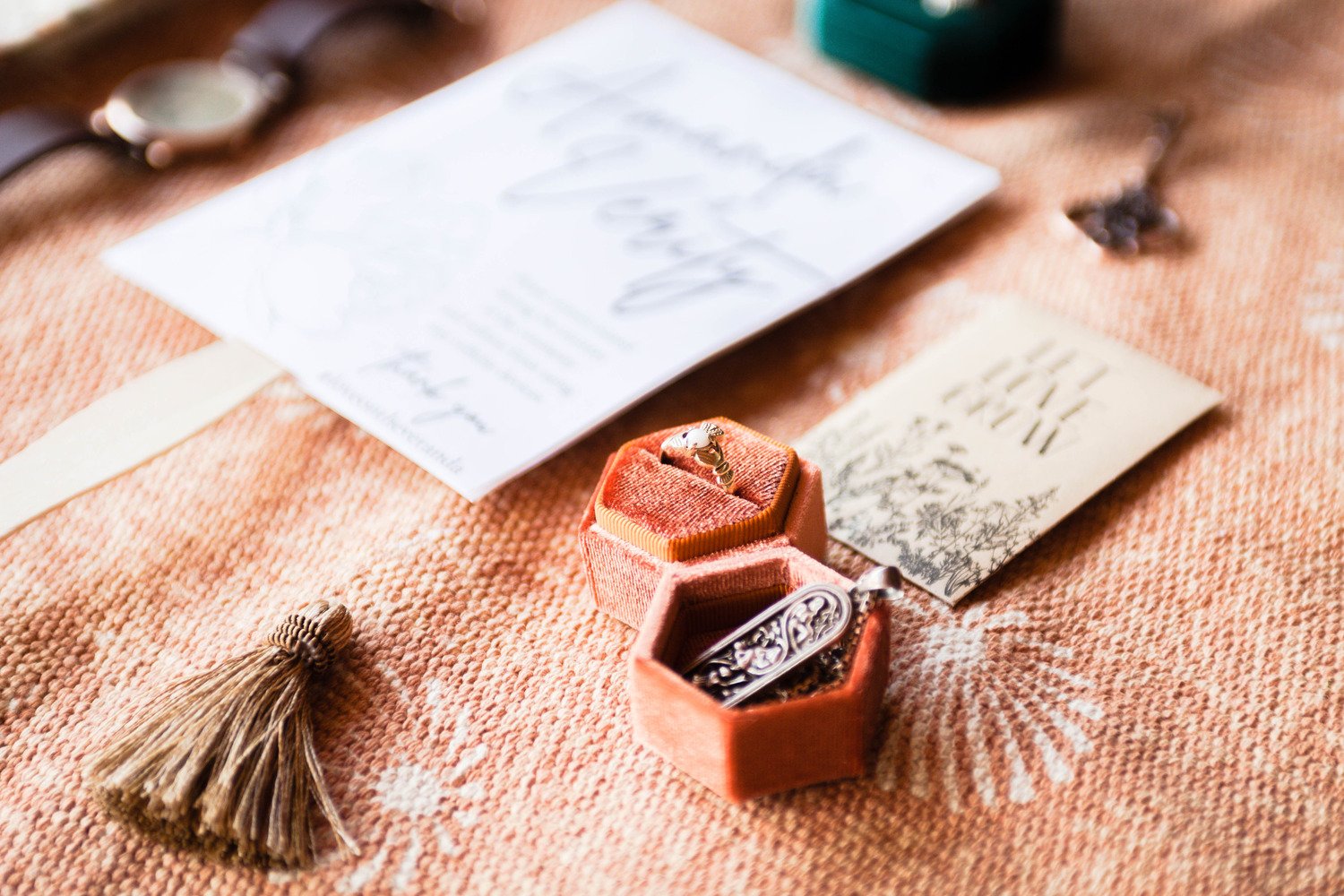 Wedding detail photos photographed on a terracotta carpet featuring wedding favors and programs with a focus on a necklace and ring in a ring box.